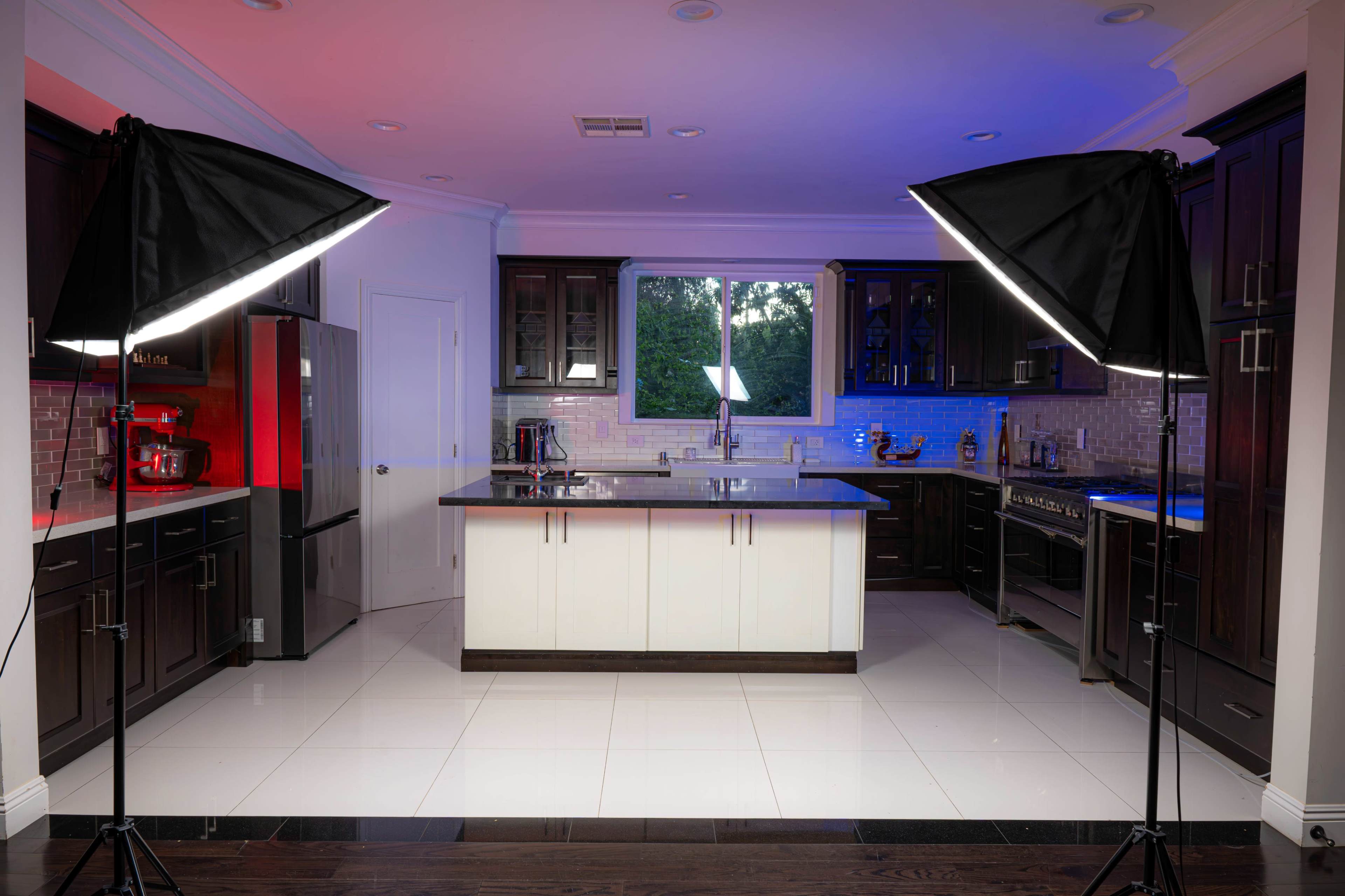 A modern kitchen is illuminated by soft box lights, featuring dark cabinetry, a center island, and stainless steel appliances.