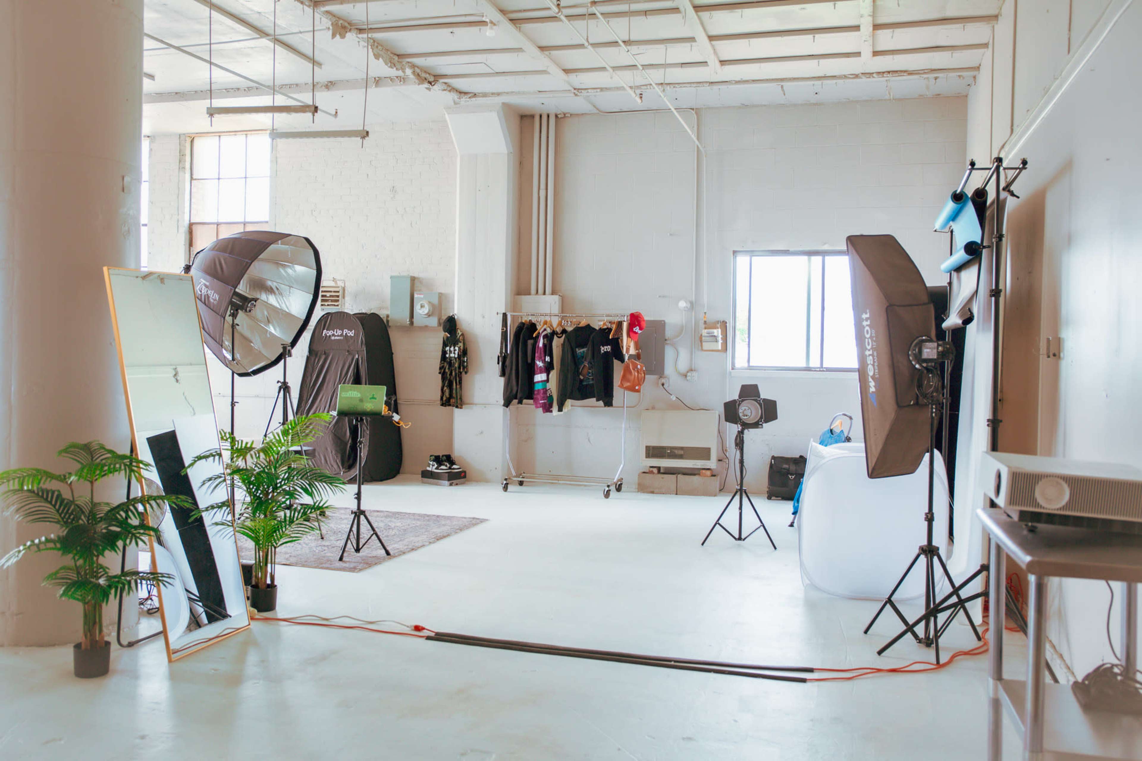 The image shows a spacious photography studio featuring softbox lights, a large mirror, clothing racks, and a projector on a stand.