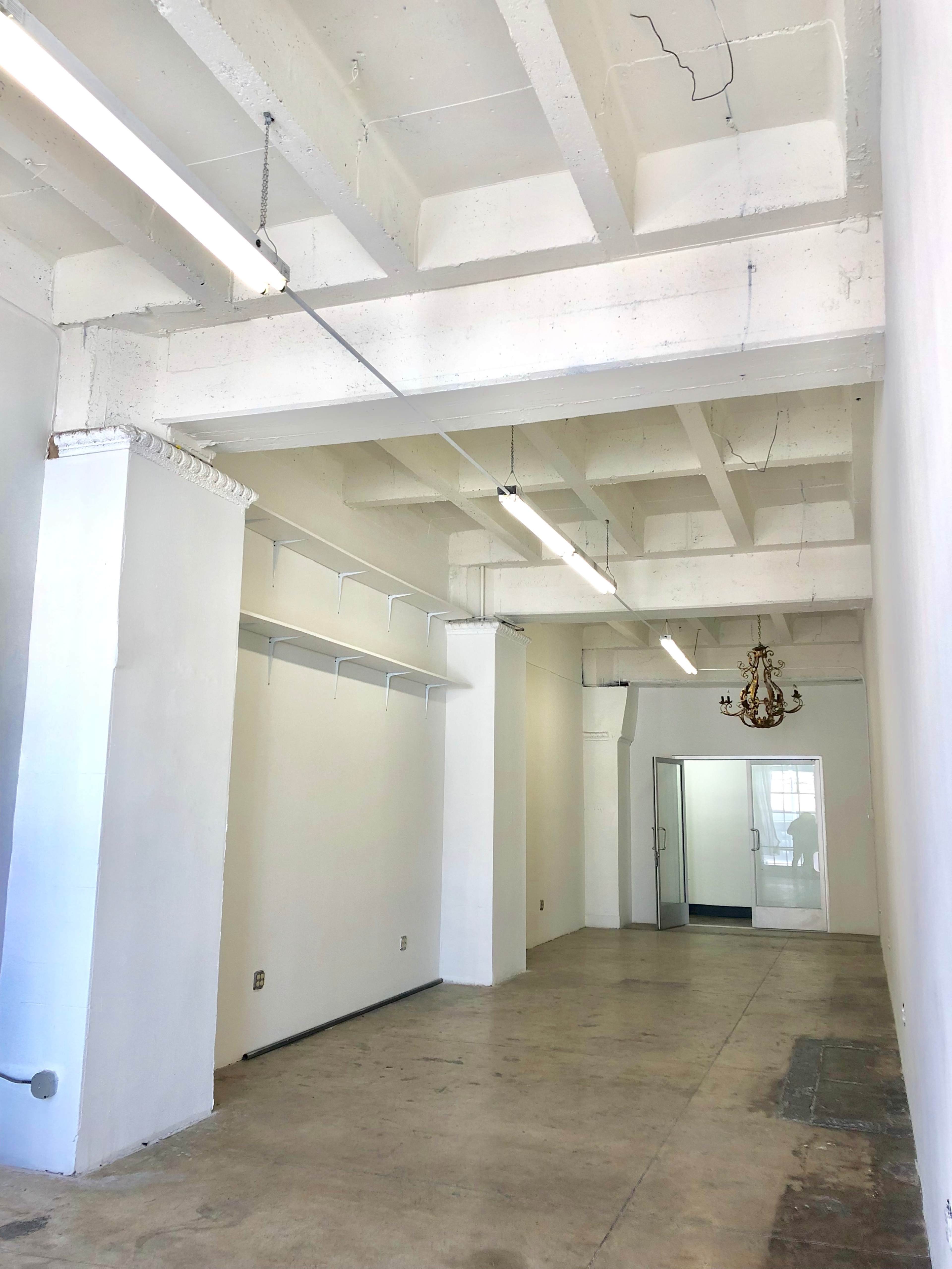 The image shows a spacious, empty room with concrete flooring, white walls, and exposed ceiling beams, featuring a chandelier and a glass door leading to another area.