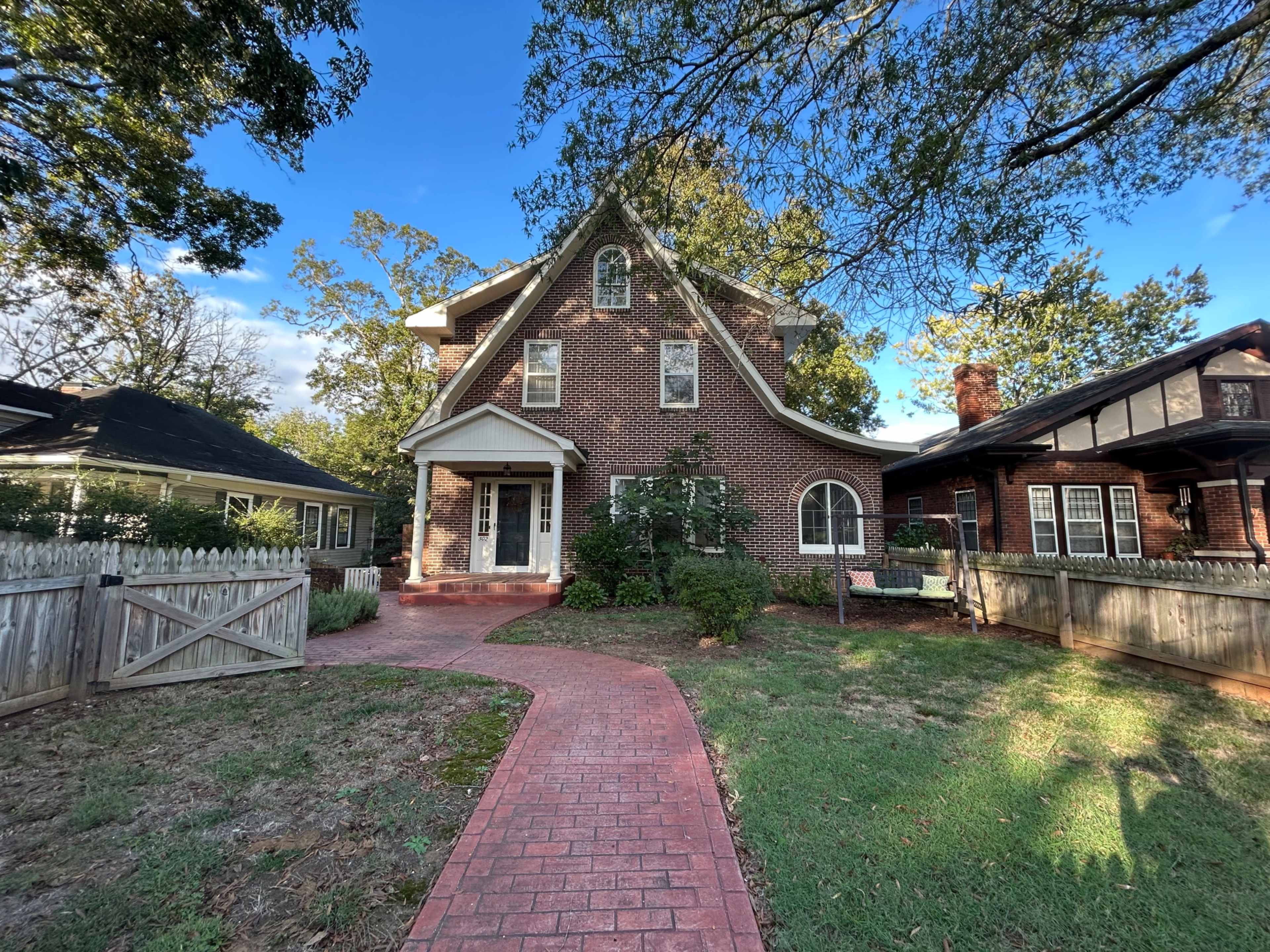 The image shows a two-story brick house with a triangular gable roof, surrounded by a green lawn and a pathway leading to the front door.