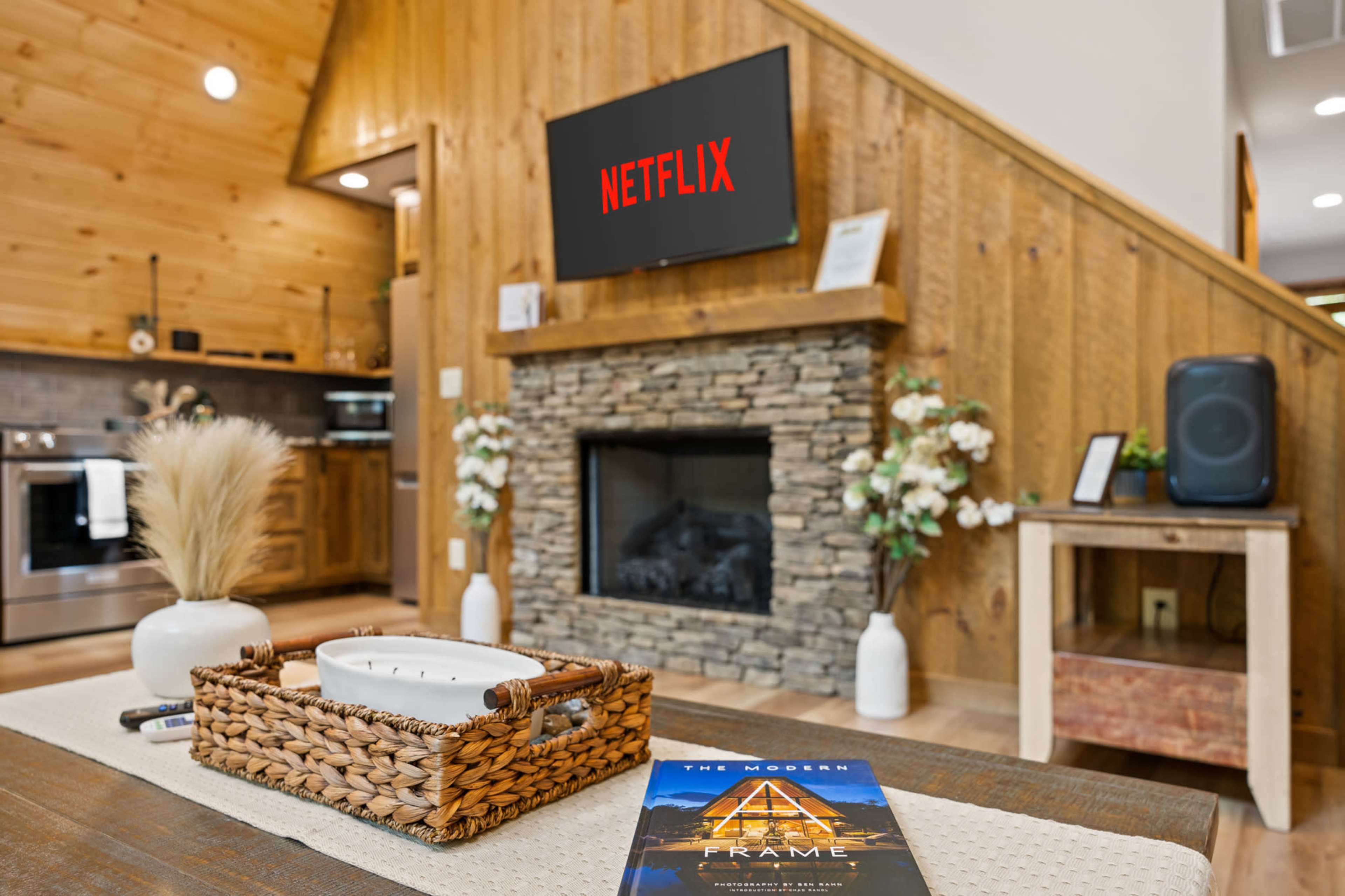 A modern living room features a stone fireplace, a television displaying the Netflix logo, and a coffee table with a woven basket and a magazine on it.