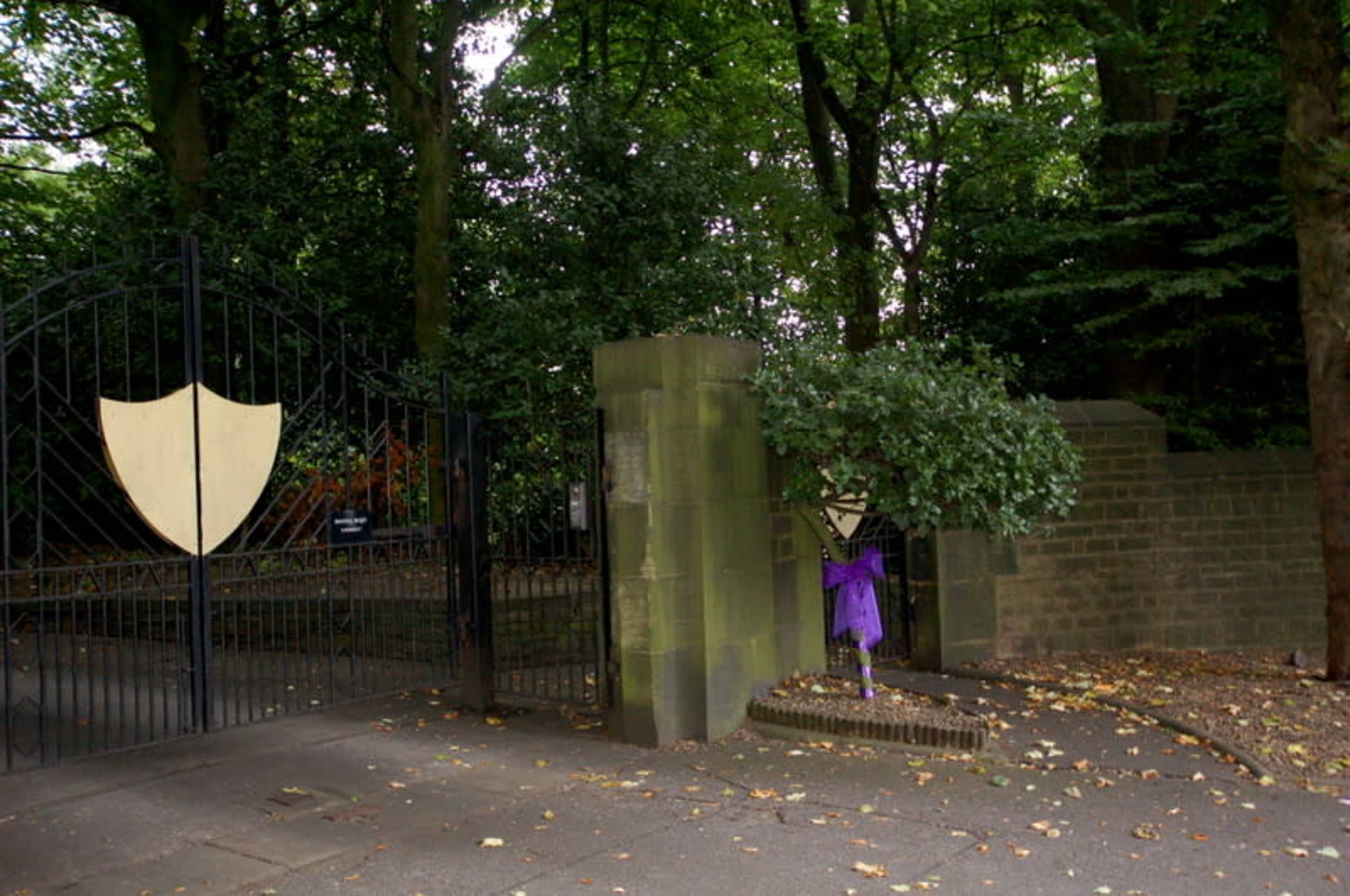 A gated entrance to a forested area, featuring tall trees, a stone wall, and decorative shields on the gate.
