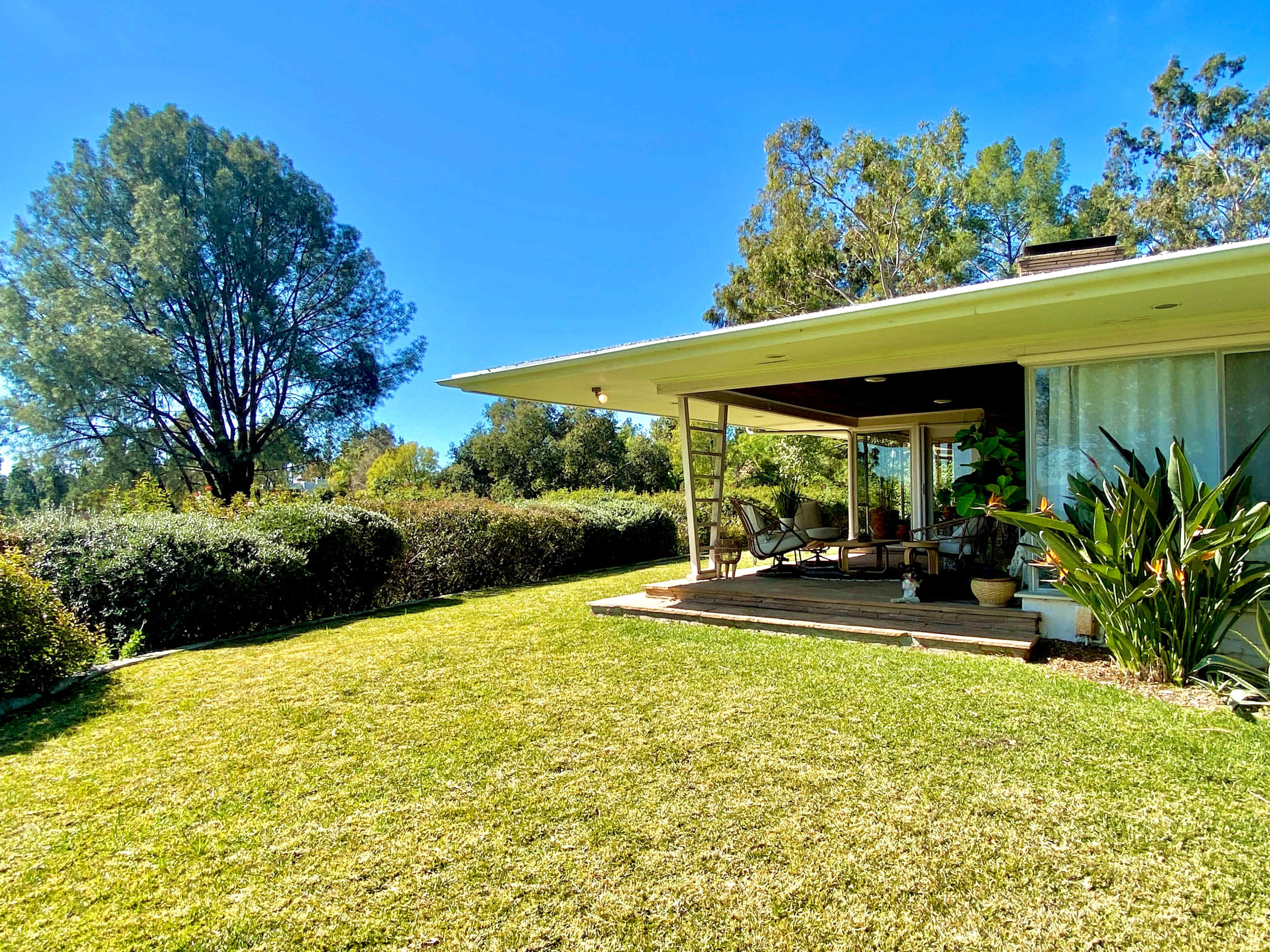 A modern house with a wooden deck and outdoor seating is surrounded by a green lawn and trees.