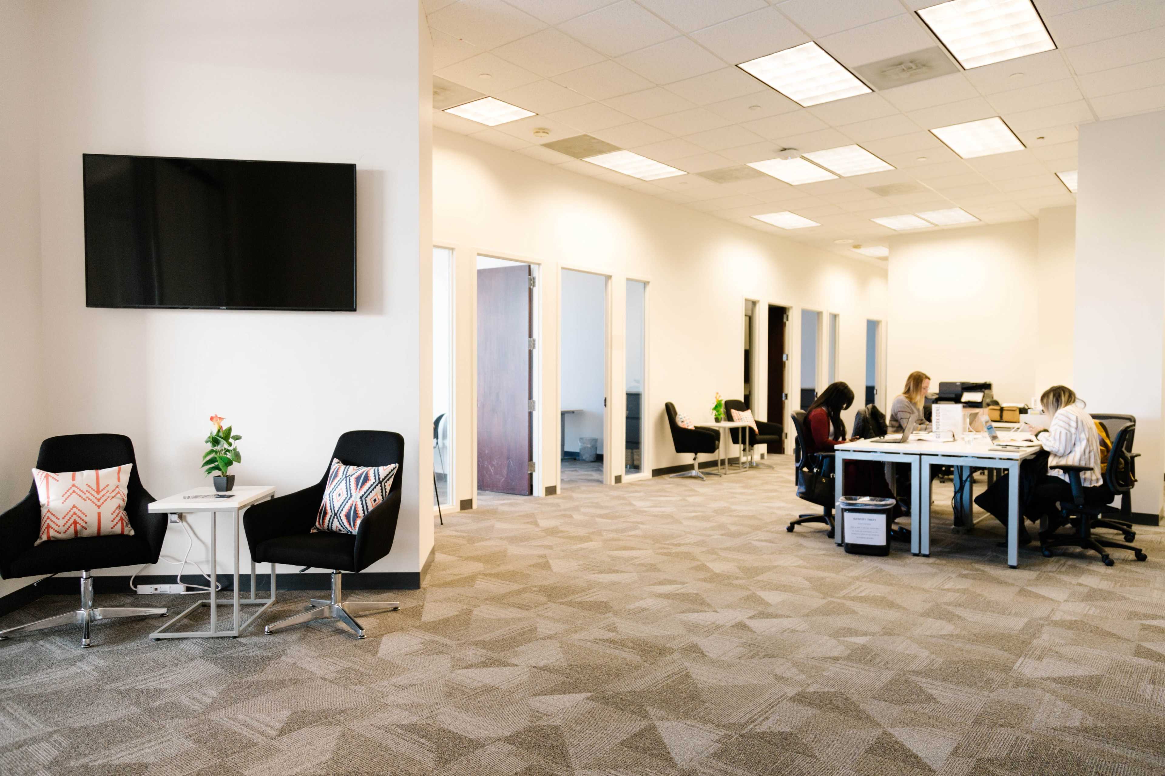 A spacious office environment with a seating area featuring chairs and a small table, and a group of people working at a table in the background.
