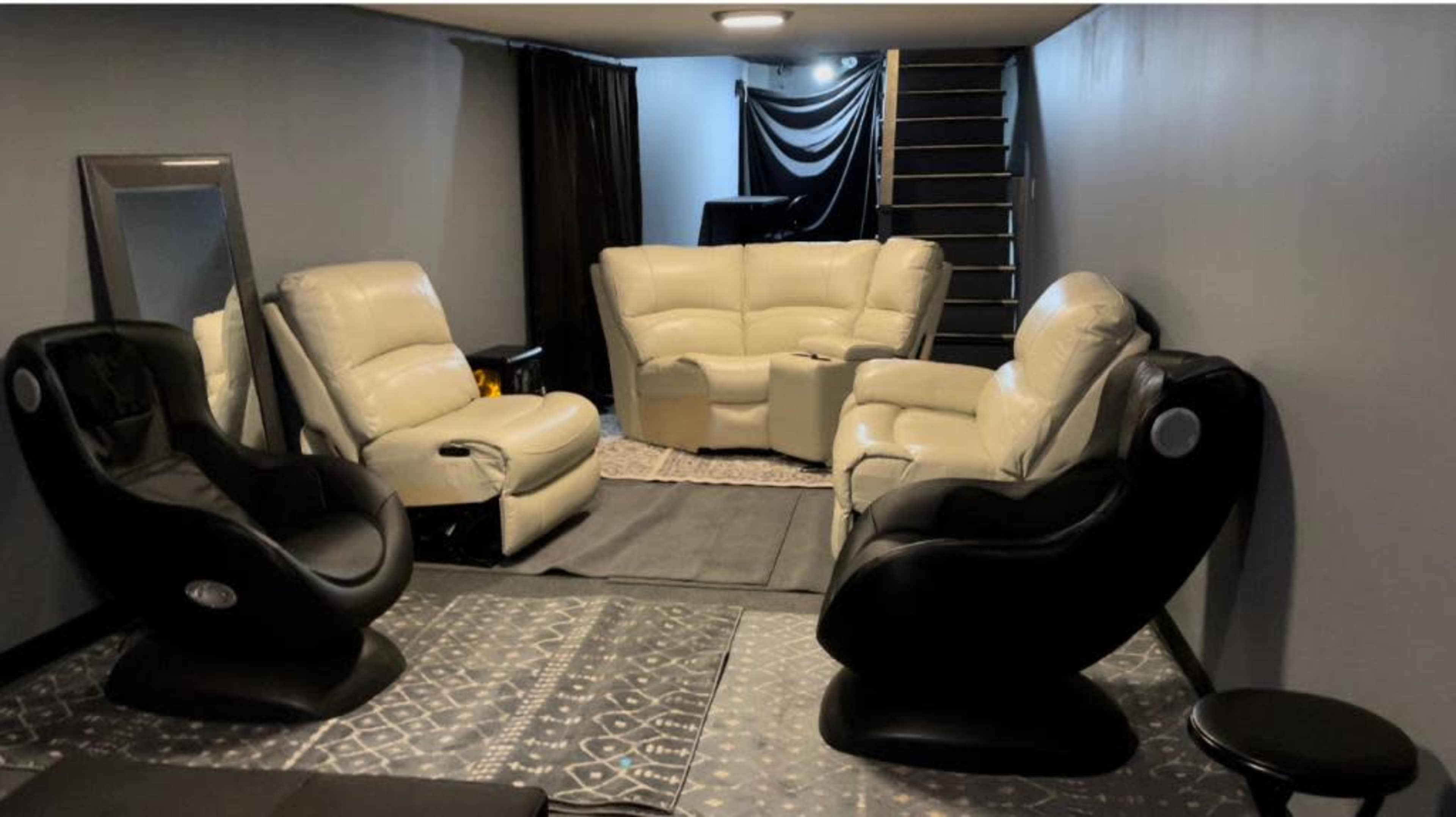 A modern, lower-level lounge area with two black and white reclining chairs, a soft sectional sofa, a small stool, and a staircase leading to a higher level.