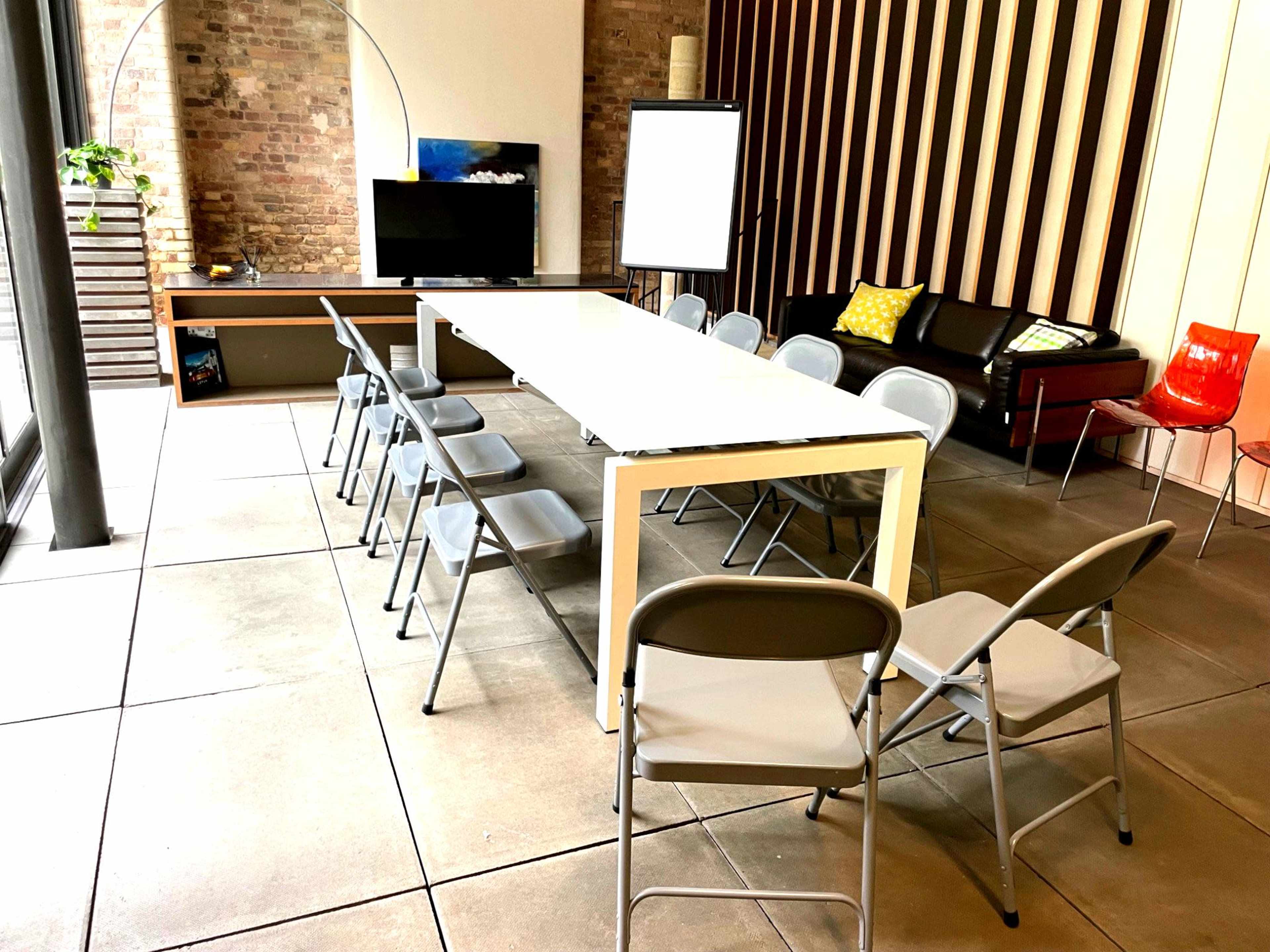 A rectangular meeting table is surrounded by gray folding chairs in a modern conference room with brick walls and a large window.