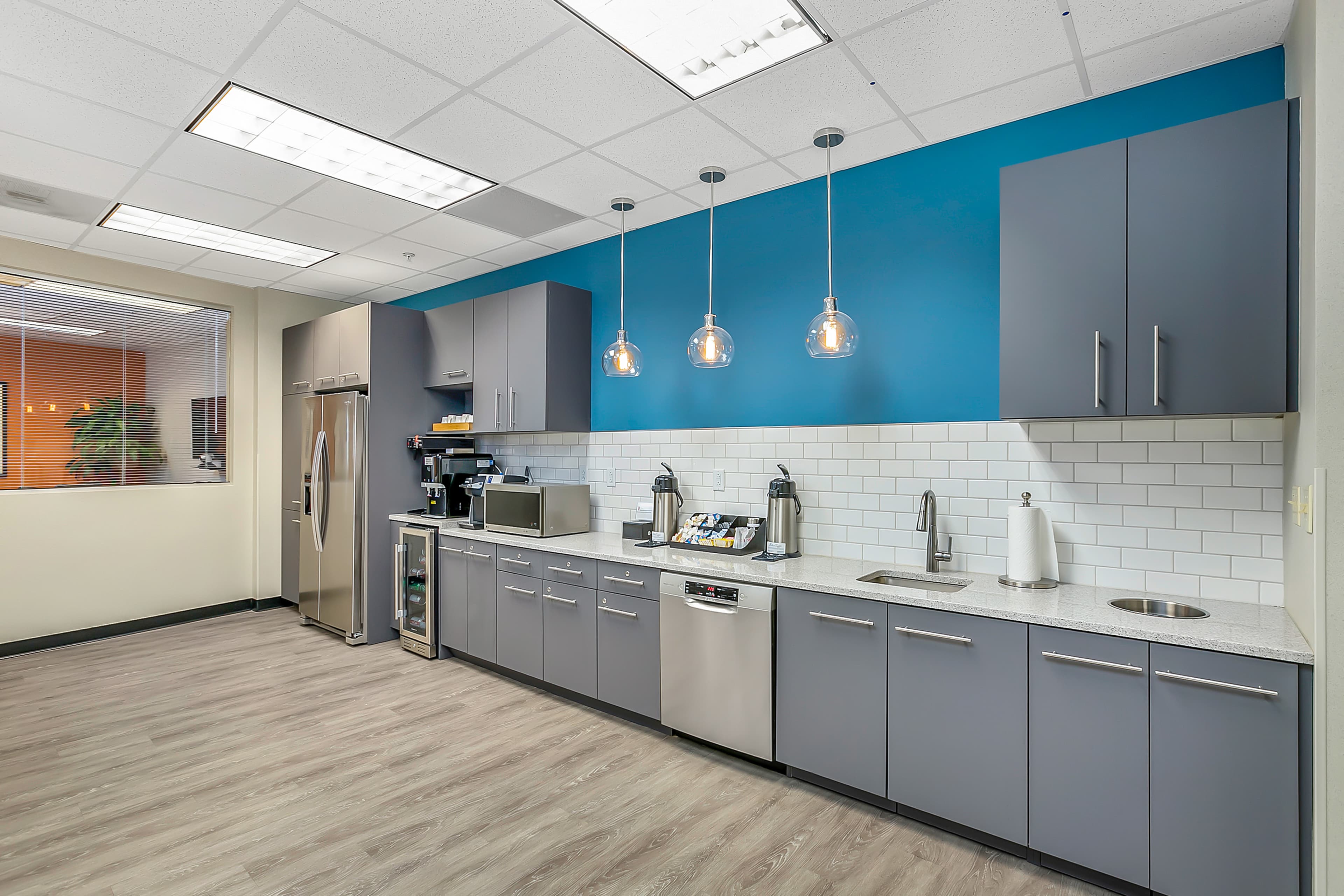 The image shows a modern kitchen with gray cabinets, a stainless steel refrigerator, and pendant lighting over a countertop with a coffee maker and various kitchen appliances.