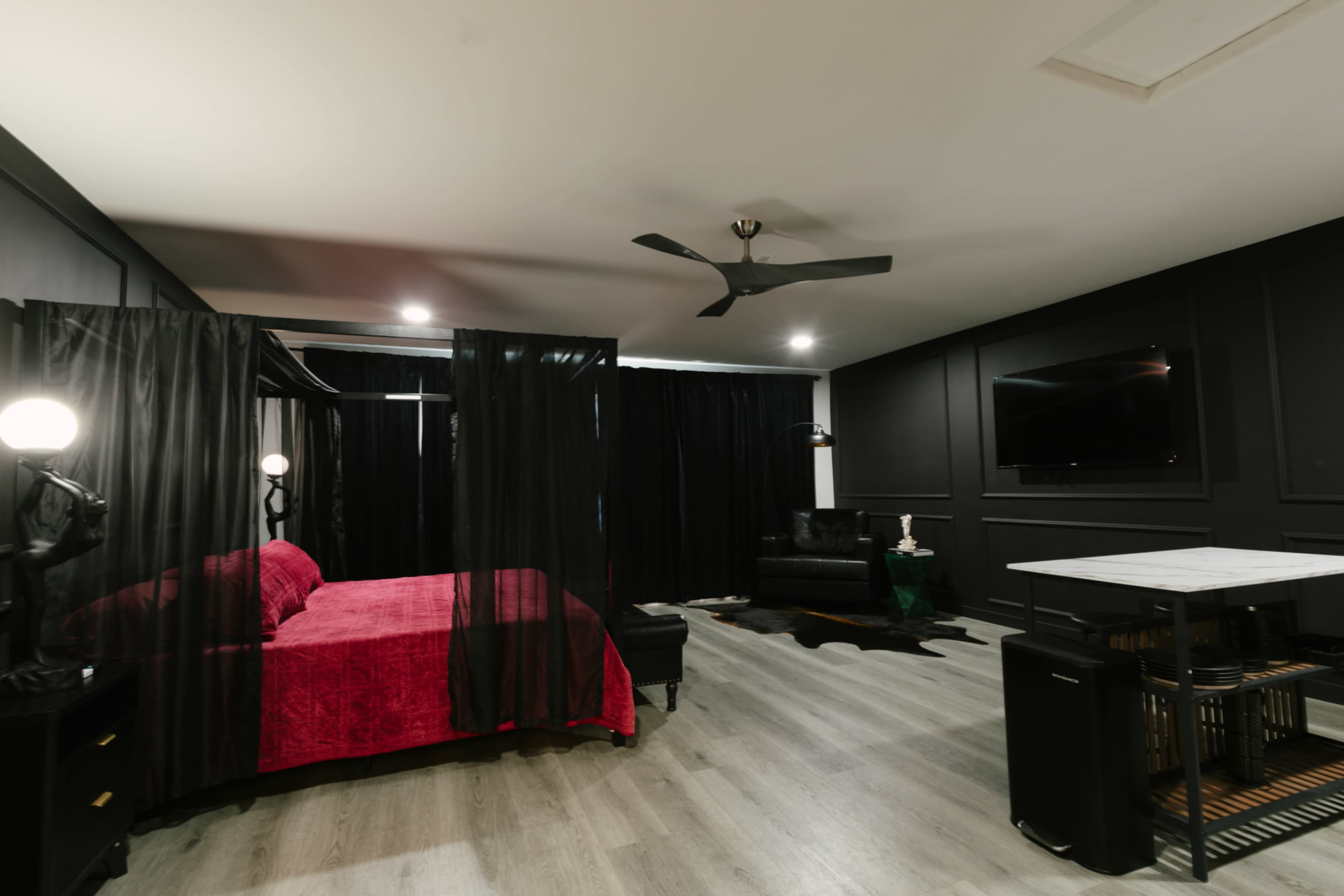 A modern bedroom features a red bed draped with black sheer curtains, accompanied by a black leather sofa, a television on the wall, and a small dining table.
