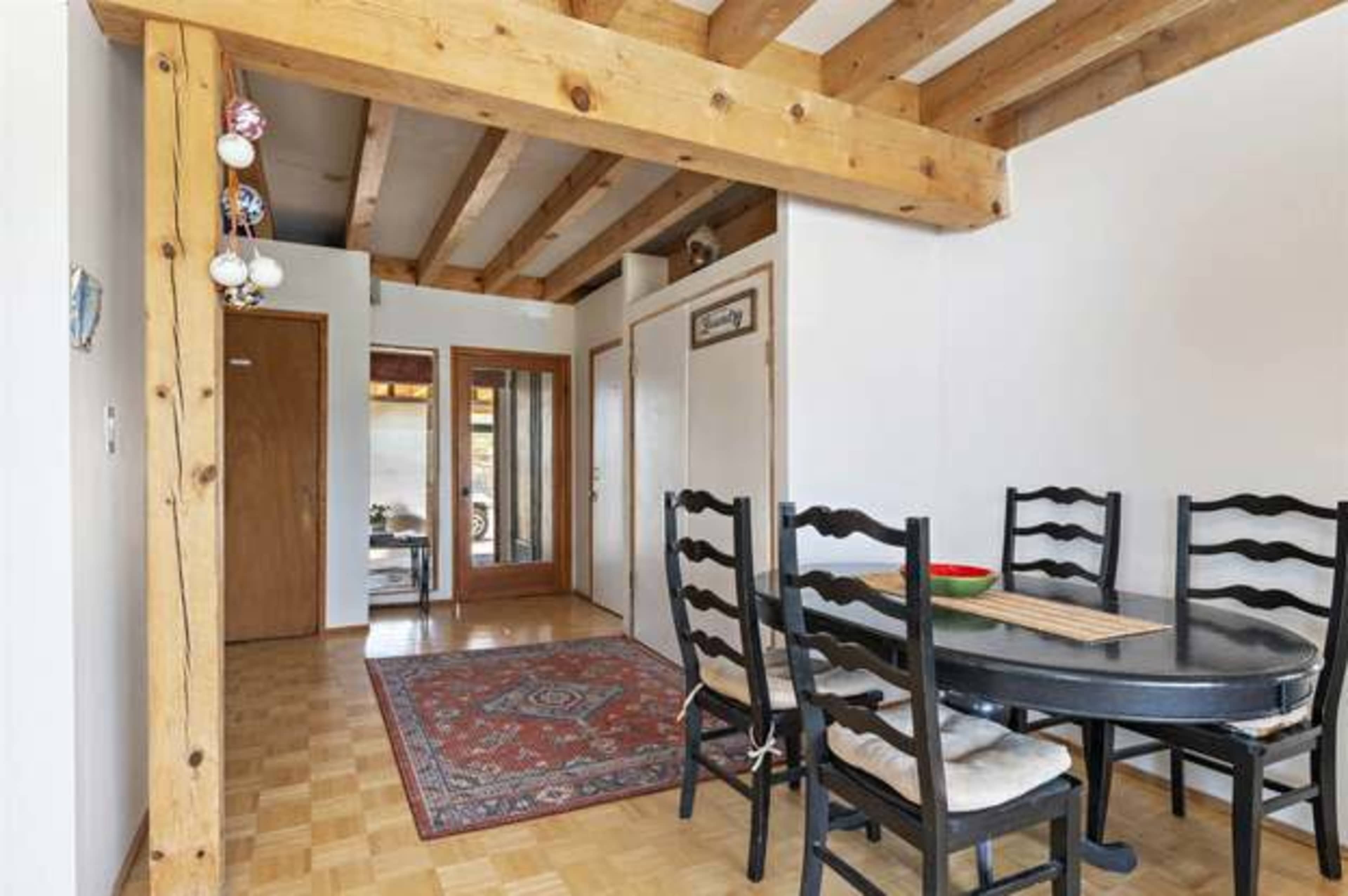 A dining area with a round black table and six chairs, set against a background of wooden beams and an adjoining hallway.