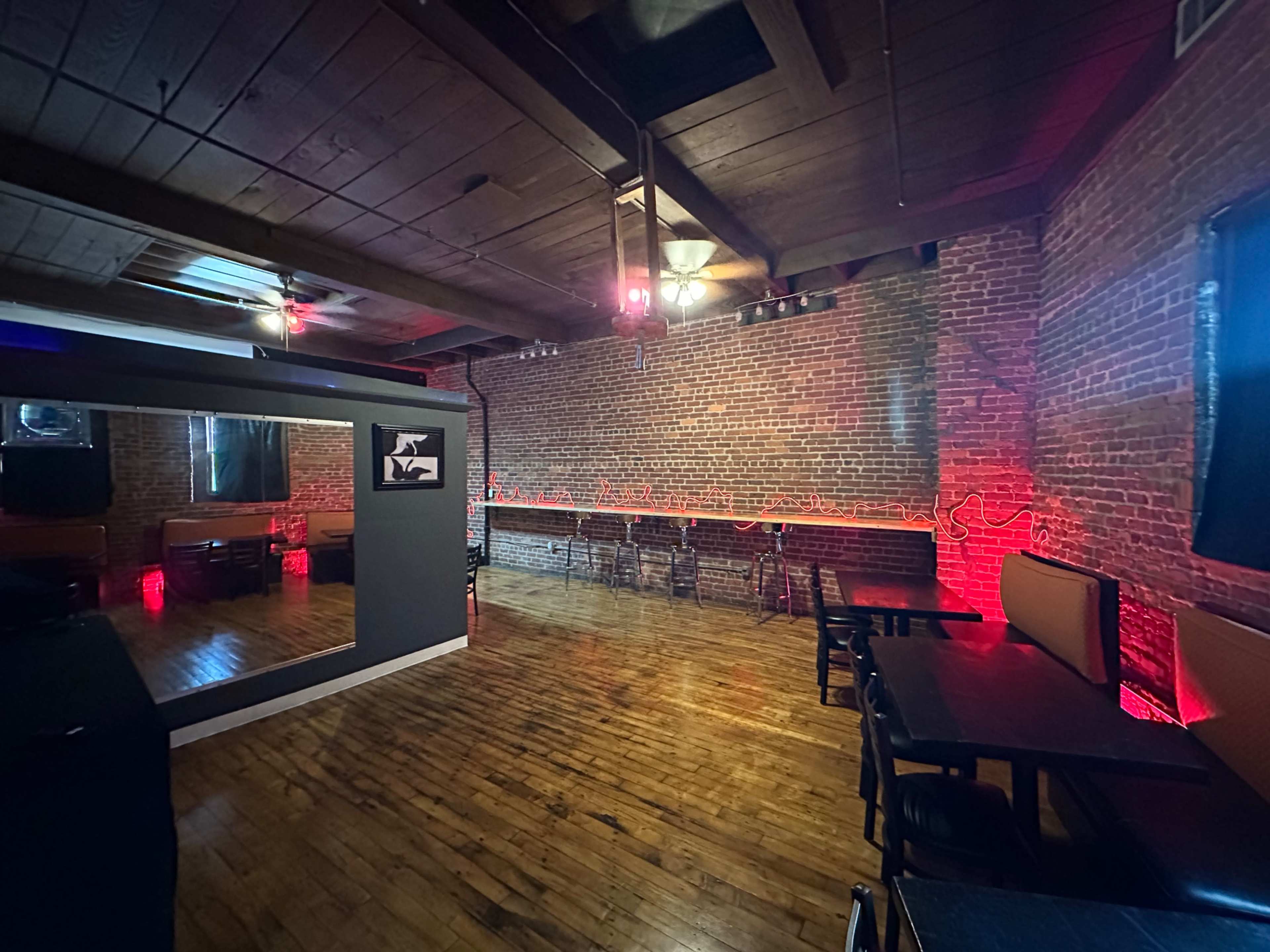 The image shows a dimly lit room with exposed brick walls, wooden floors, and several empty black tables and chairs arranged along the sides.
