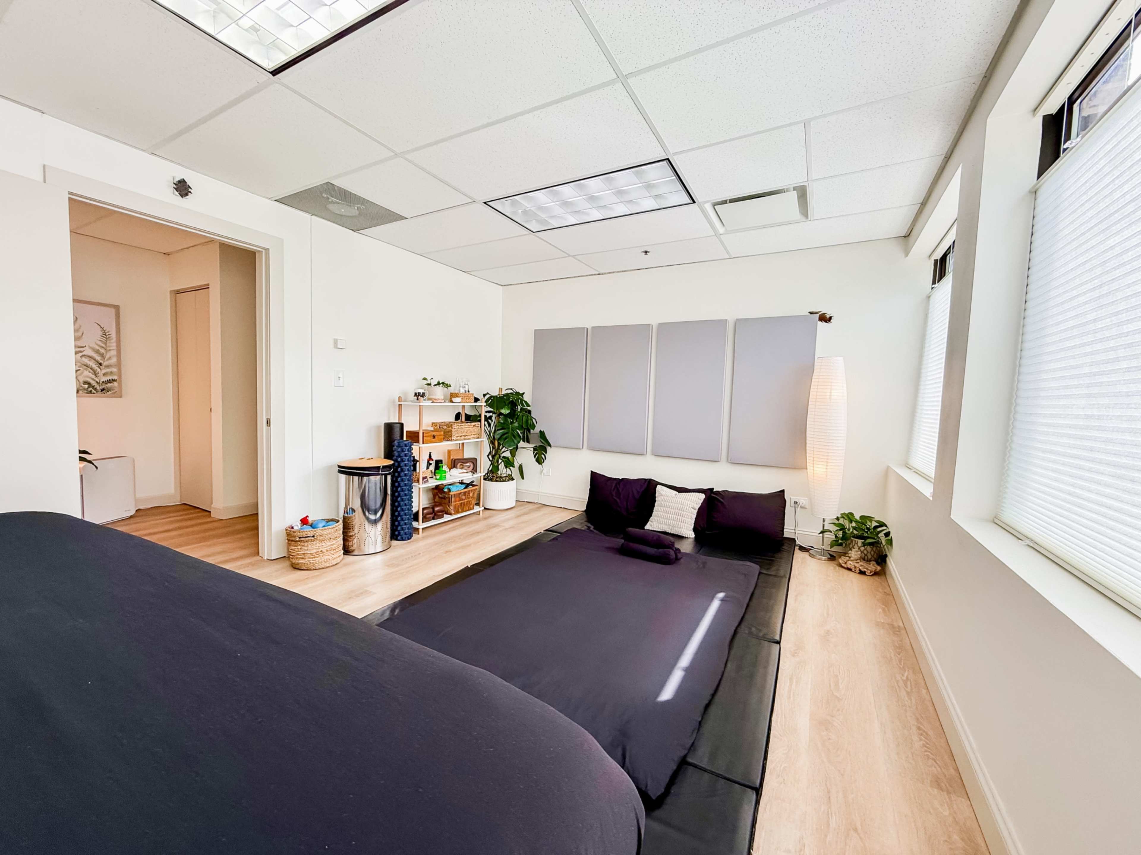 The image shows a minimalist bedroom with a dark bedding setup, a small seating area with plants, and wall-mounted panels for decoration.