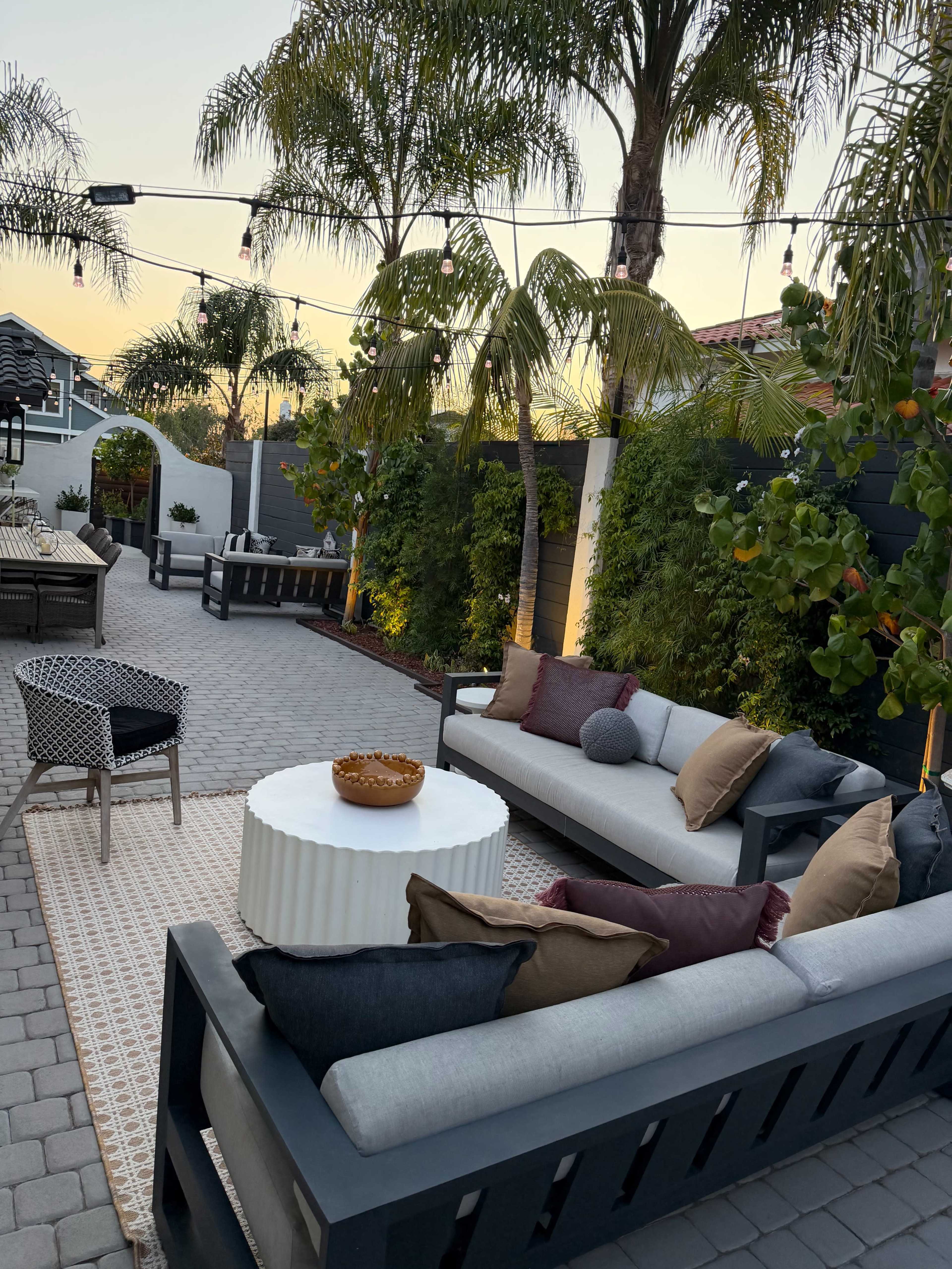 The image shows a patio area with several seating arrangements, including a gray couch with cushions, a round table, and decorative plants under string lights at sunset.