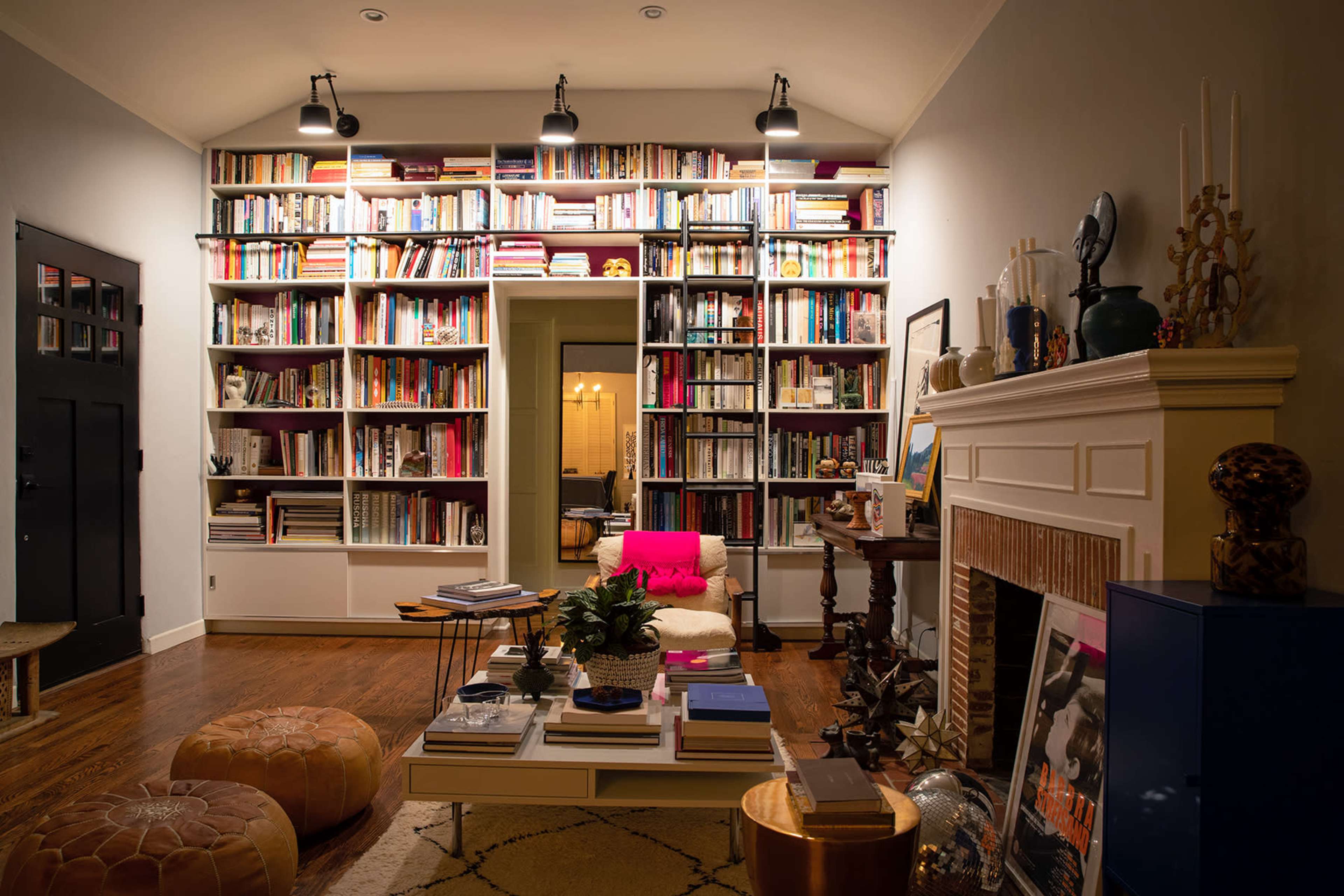 A cozy living room with a large bookshelf filled with books, a white chair, a coffee table stacked with books, and decorative objects around a fireplace.