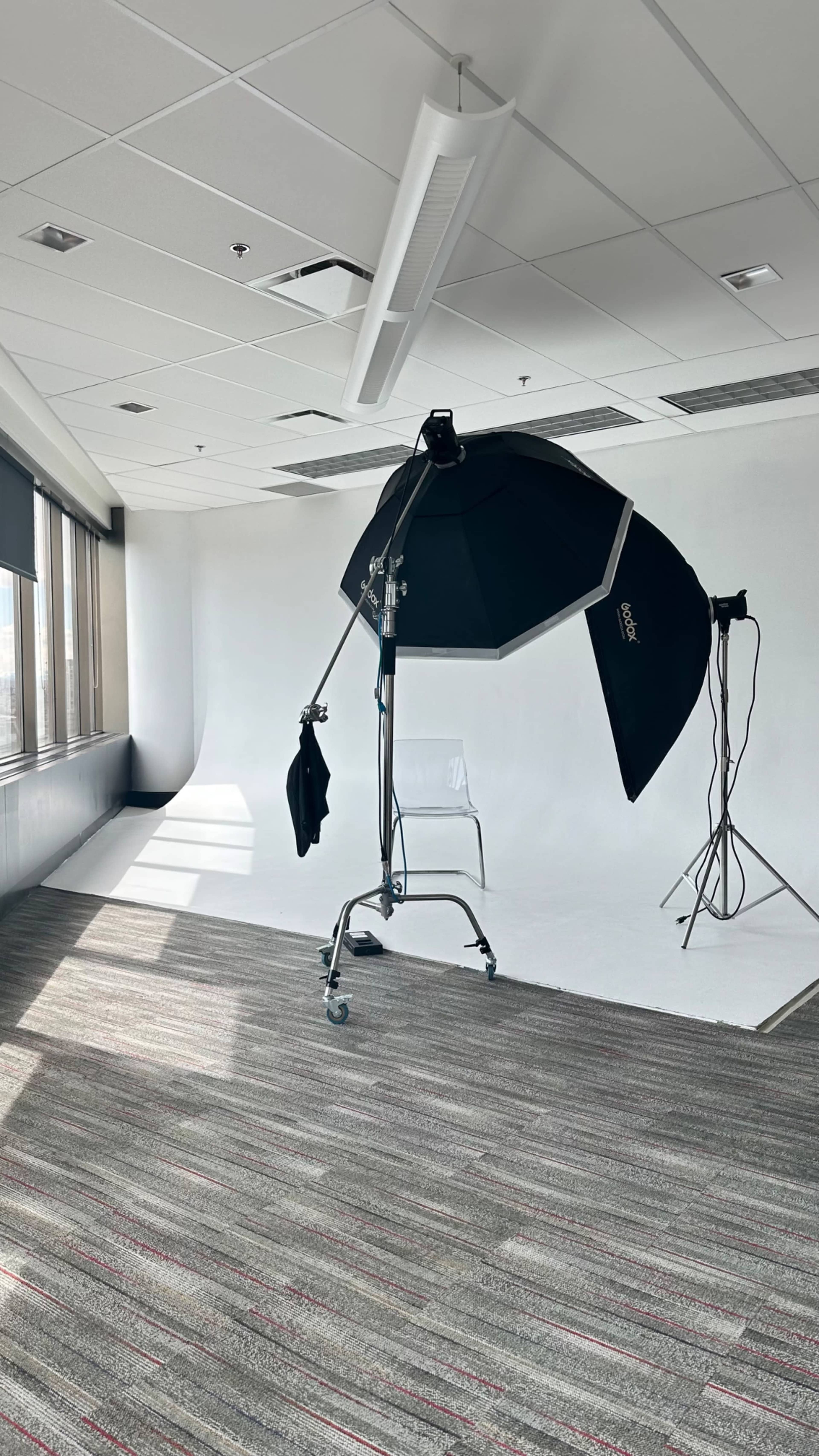 A photography studio setup features a large softbox on a stand and a backdrop against a white wall with a patterned carpet.