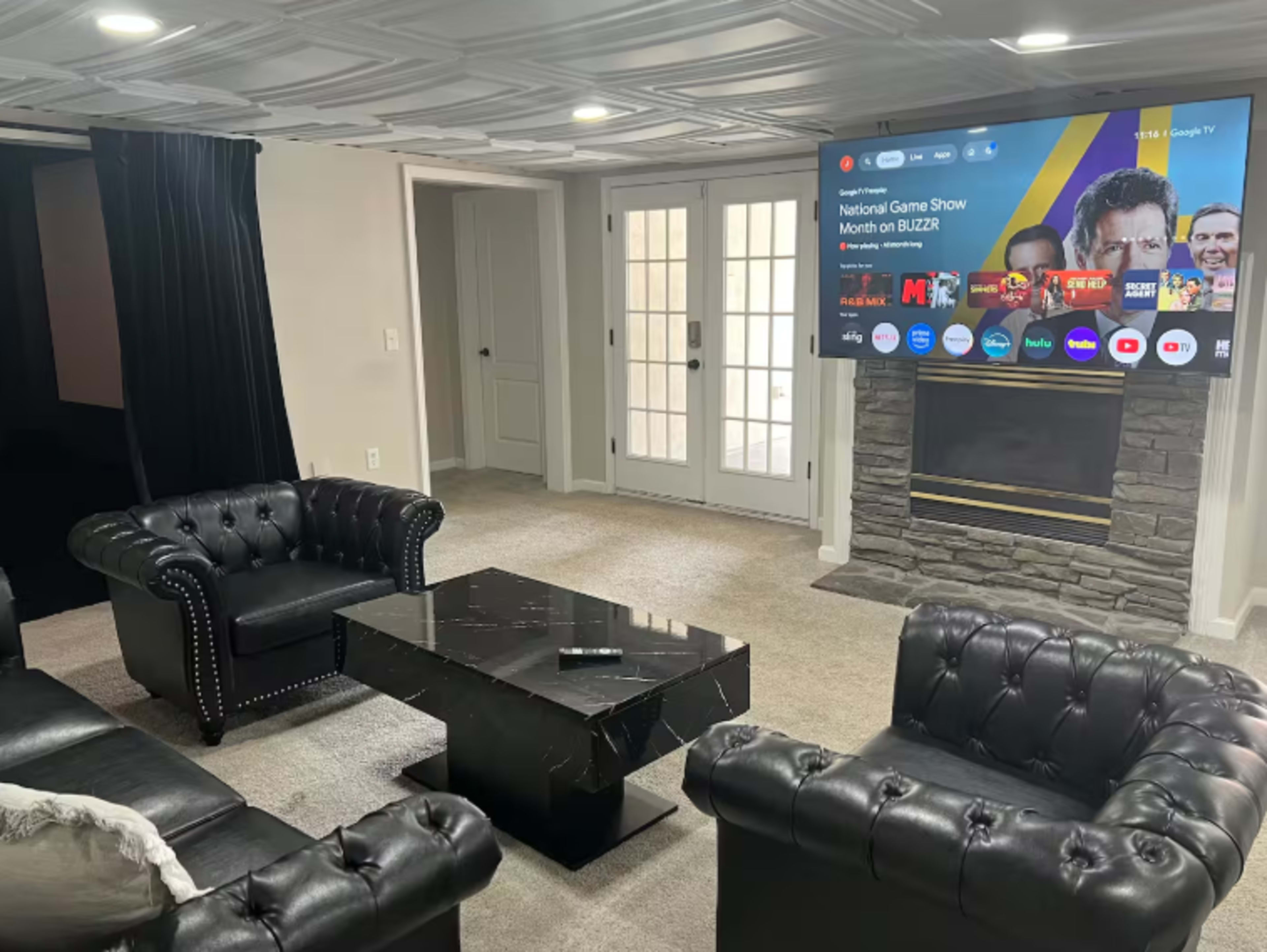 The image shows a modern living room with two black leather chairs, a glass coffee table in the center, and a large television mounted on the wall above a stone fireplace.