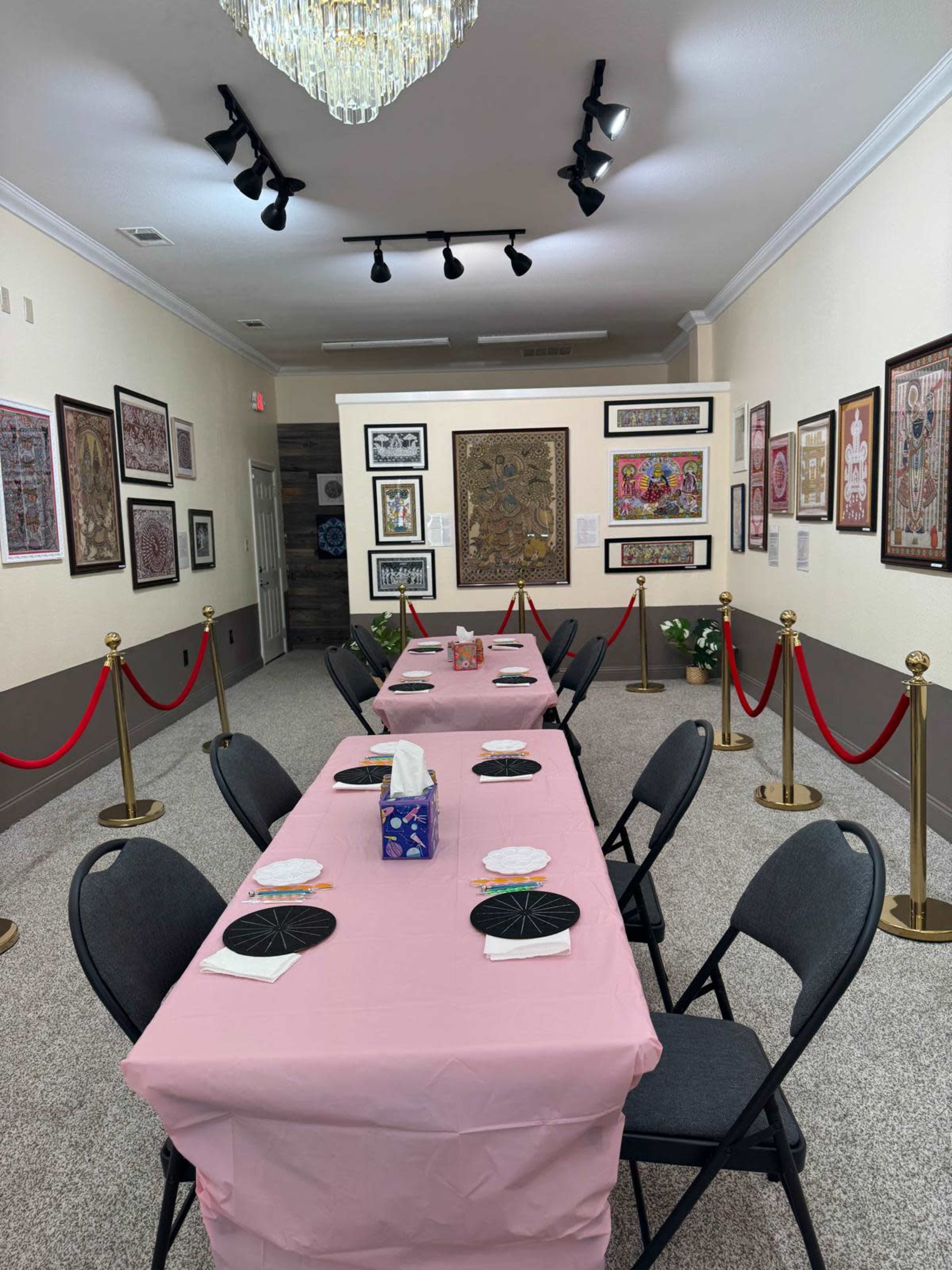 The image shows a decorated room with a long table set with pink tablecloths and black circular place settings, flanked by artwork on the walls and a red rope barrier.