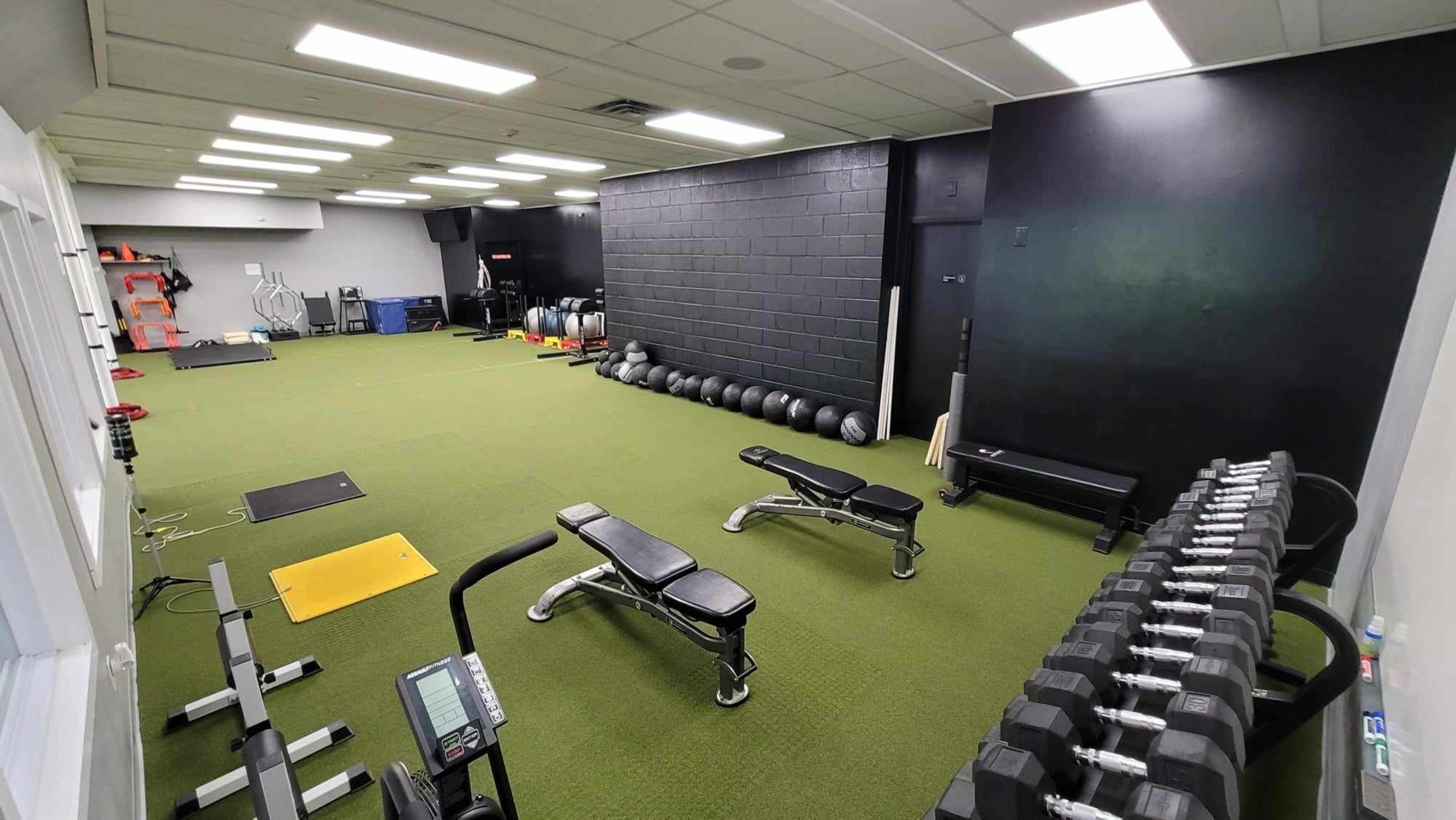 An indoor gym space with green flooring, various exercise equipment, including benches, dumbbells, and medicine balls, arranged along the walls.