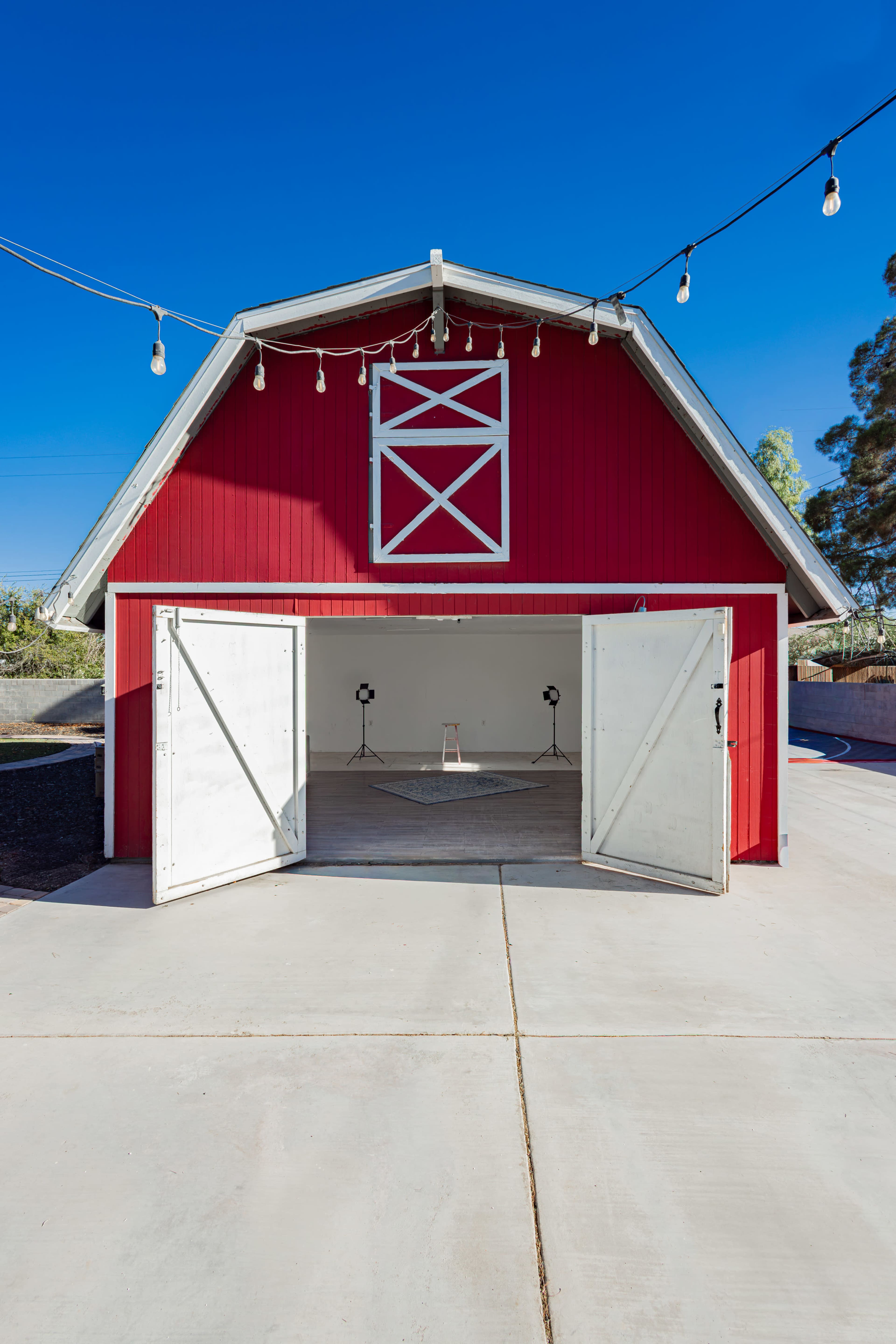 A red barn with white doors is open, showcasing a simple interior and string lights overhead against a clear blue sky.