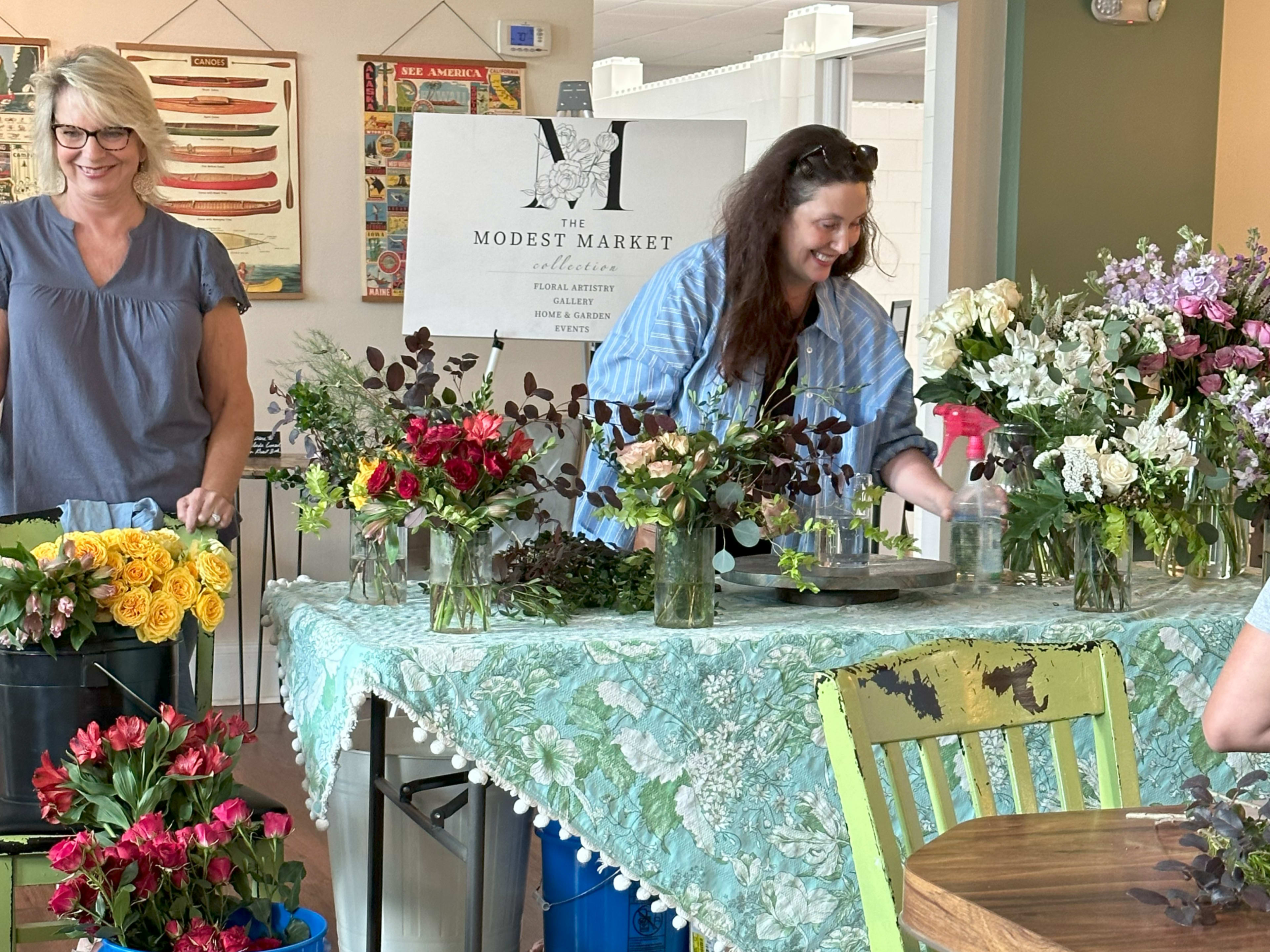 Two women are arranging flowers at a table decorated with a green cloth and surrounded by vases filled with various blooms.