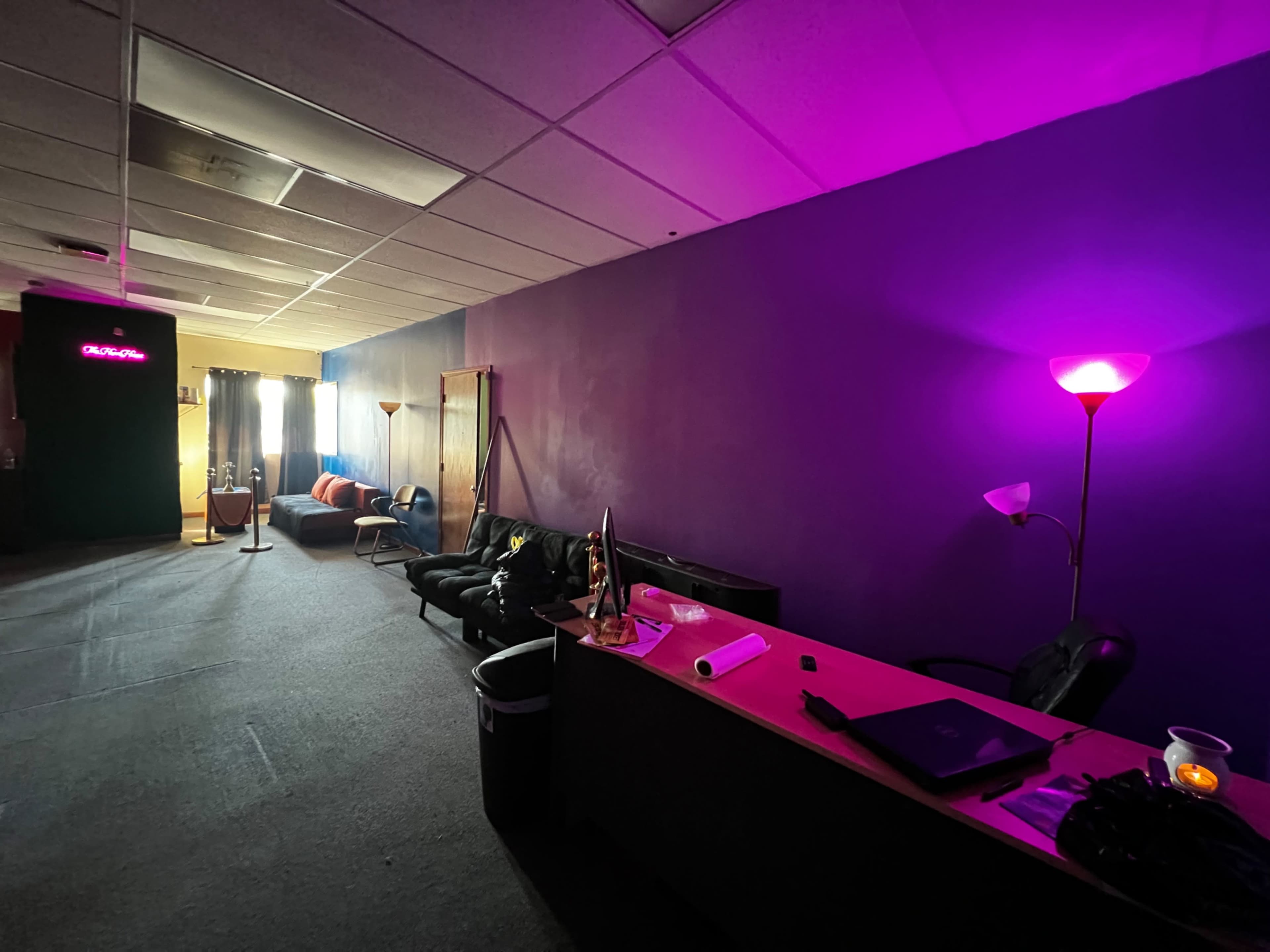 A brightly colored room with a dark purple wall, a black couch, a desk with items on it, and a lamp providing light on the right side.