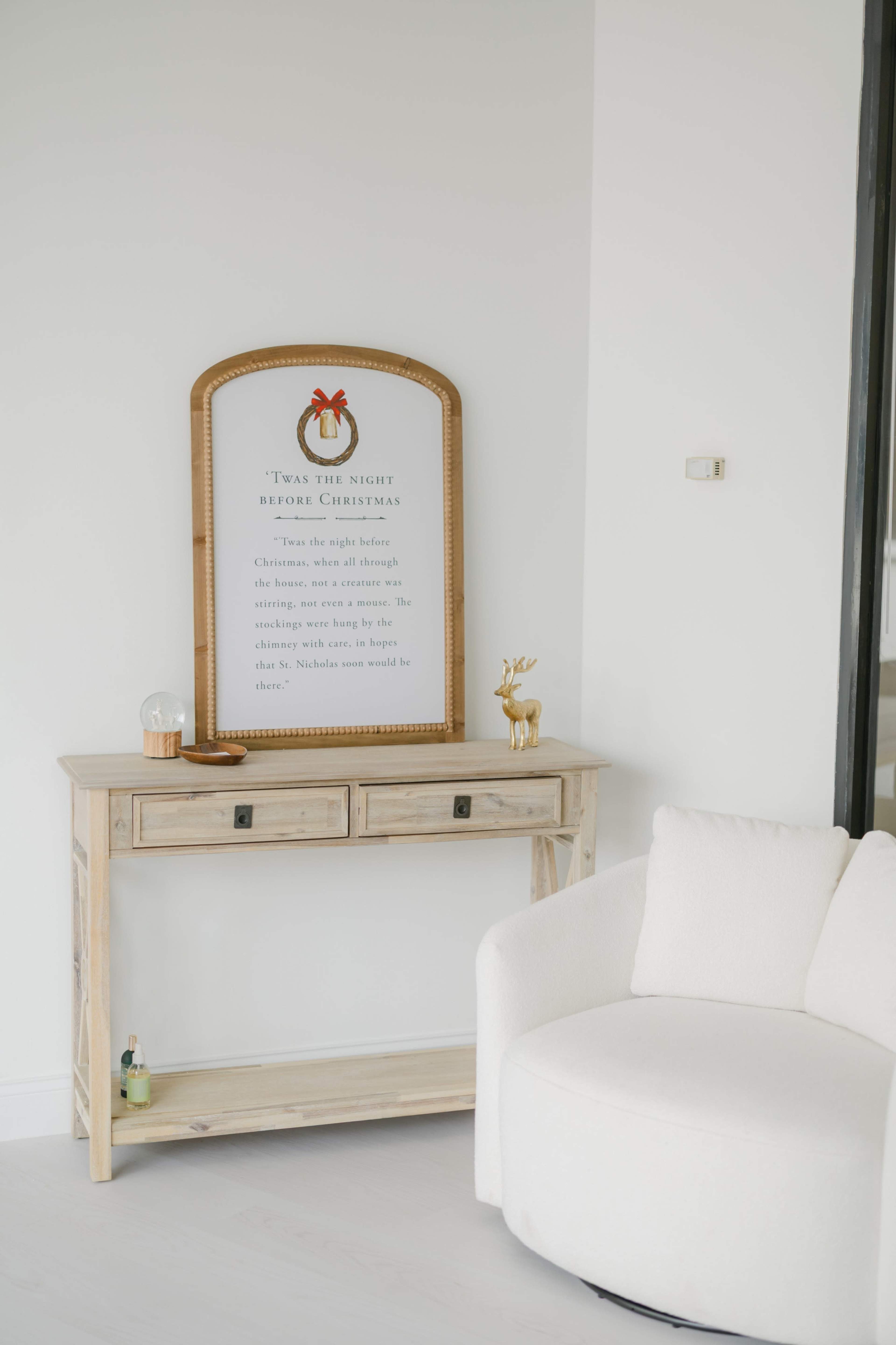 A wooden console table with two drawers stands against a white wall, featuring a decorative mirror with a Christmas-themed text above it, alongside a small ornament and a decorative reindeer figurine.