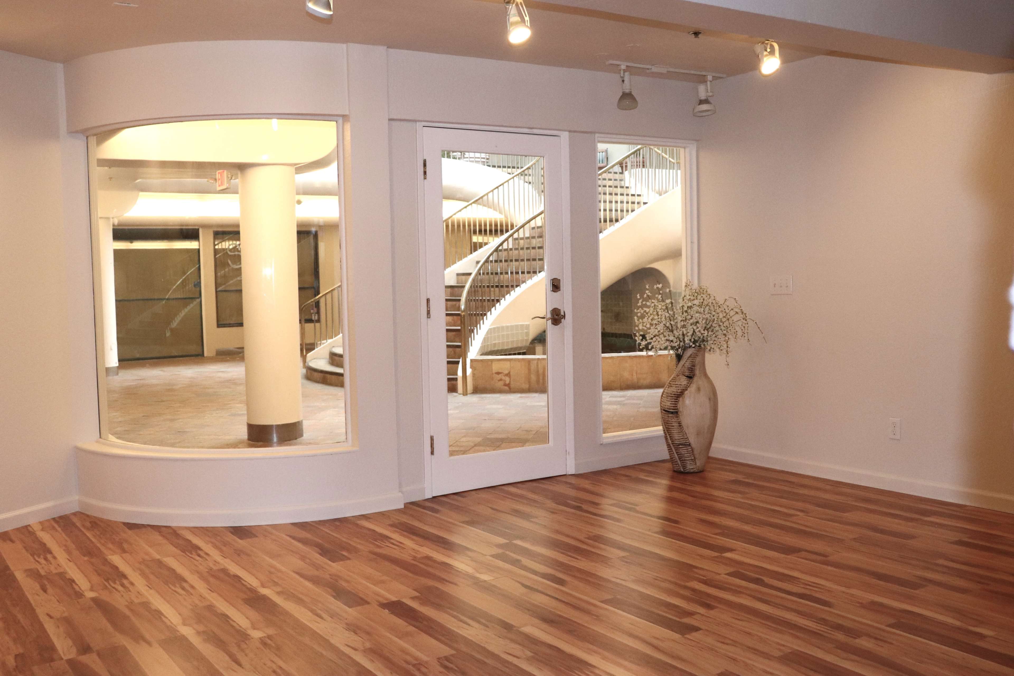 A modern interior room with wooden flooring, featuring a large window, double doors, and a staircase visible through another window.