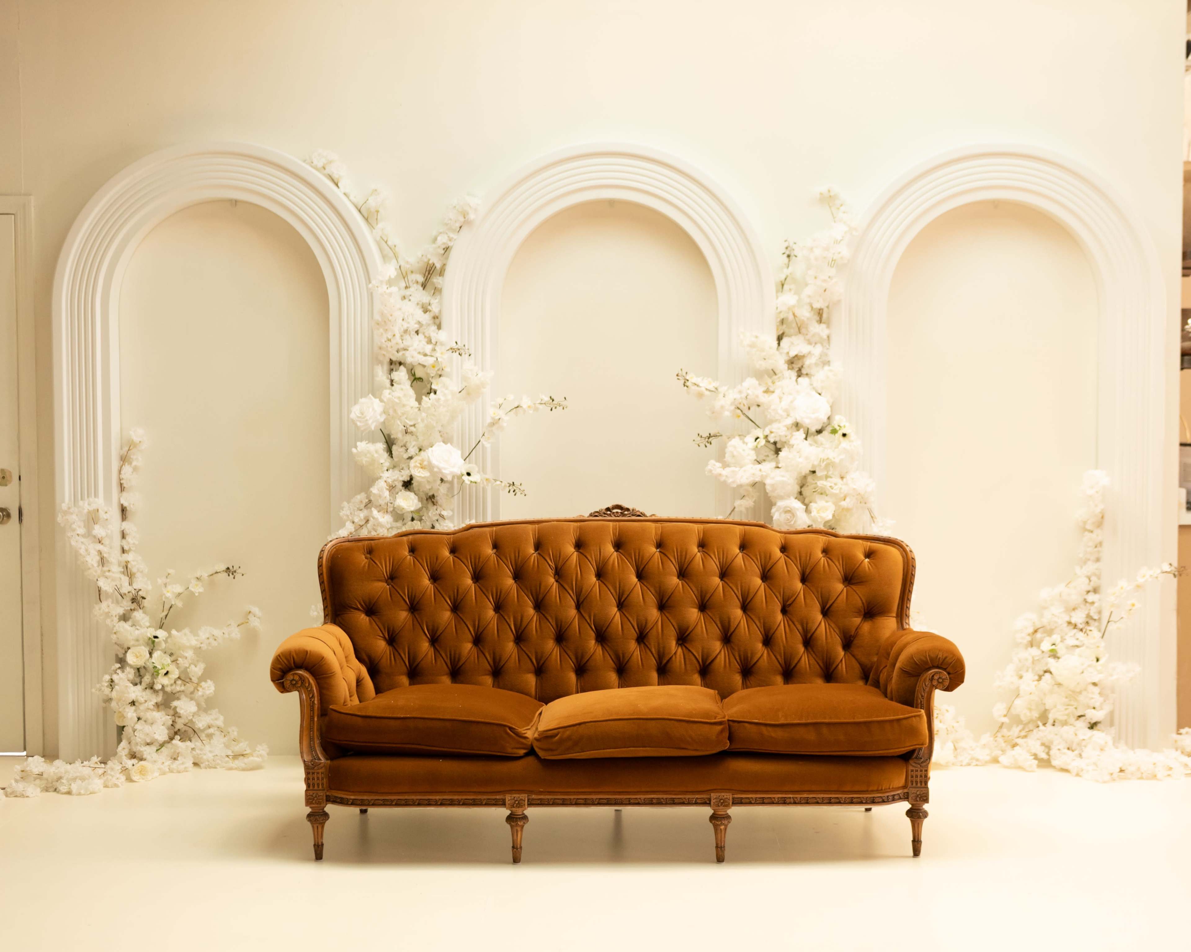A tufted vintage sofa is centered against a plain wall adorned with three arch-shaped decorations and white floral arrangements.