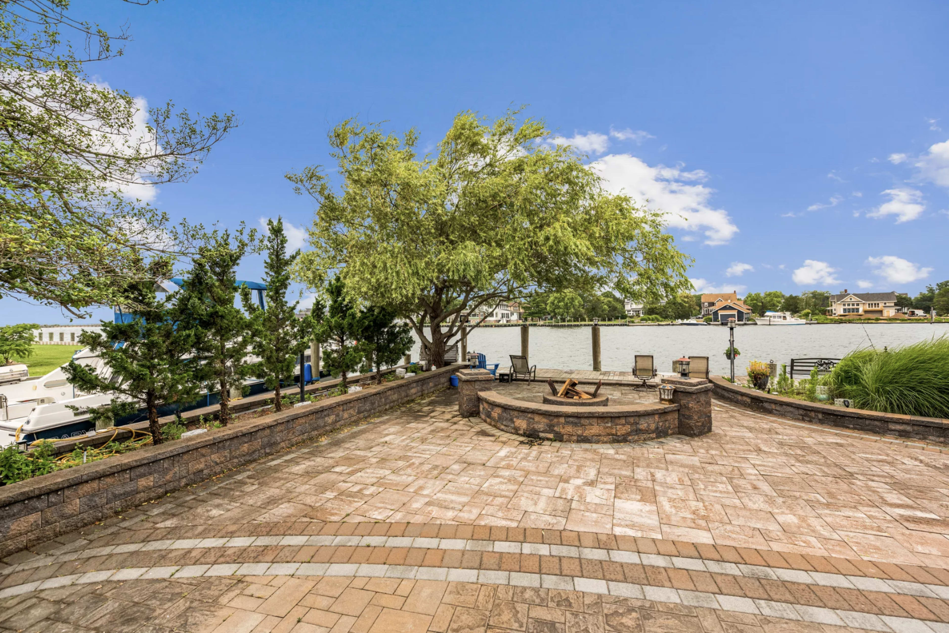 A paved outdoor patio with a circular fire pit surrounded by seating, overlooking a waterfront area with boats moored nearby.