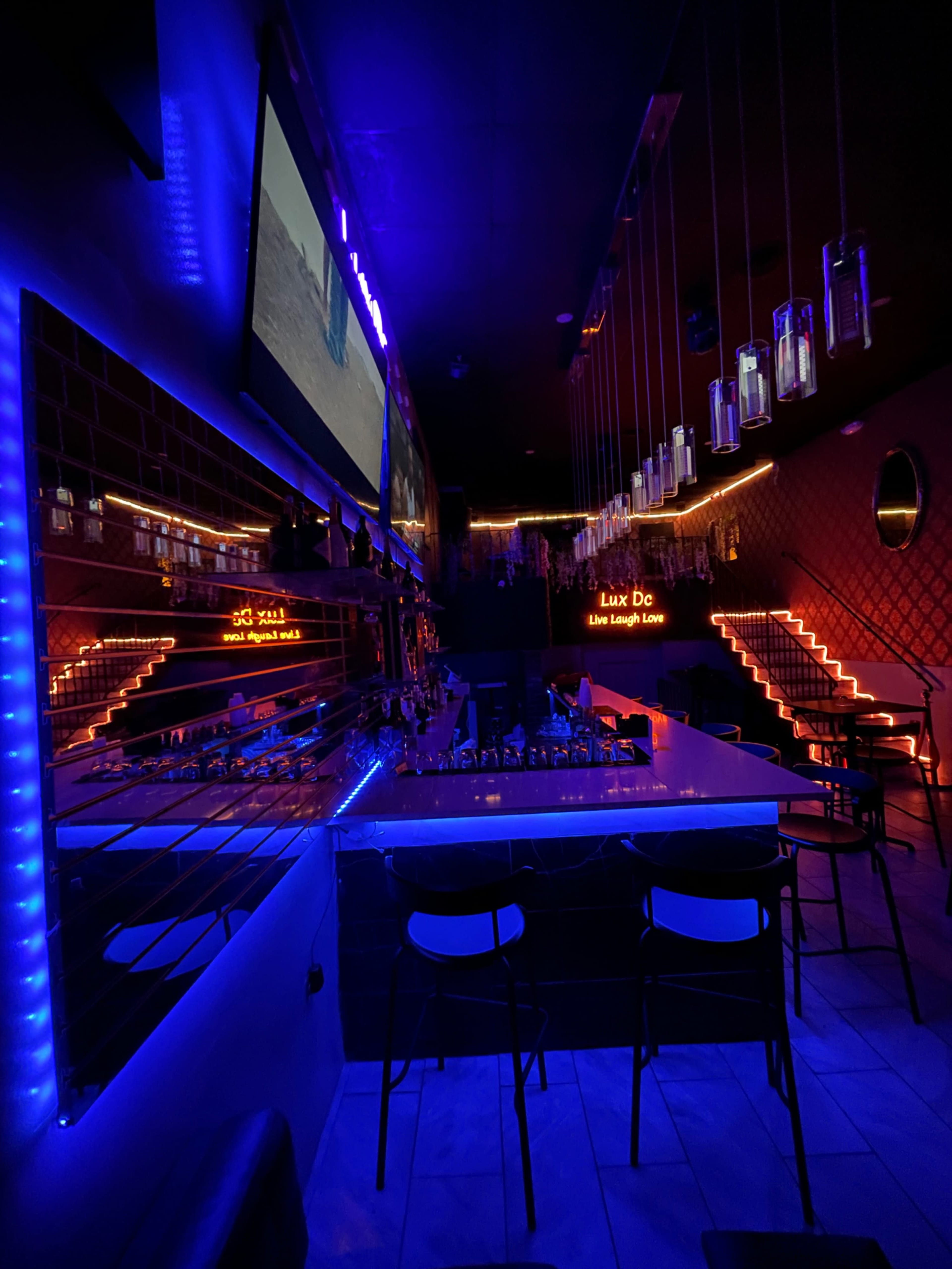 The image shows a dimly lit bar with blue and red neon lighting, featuring a sleek countertop and mirrored walls, along with illuminated stairs in the background.