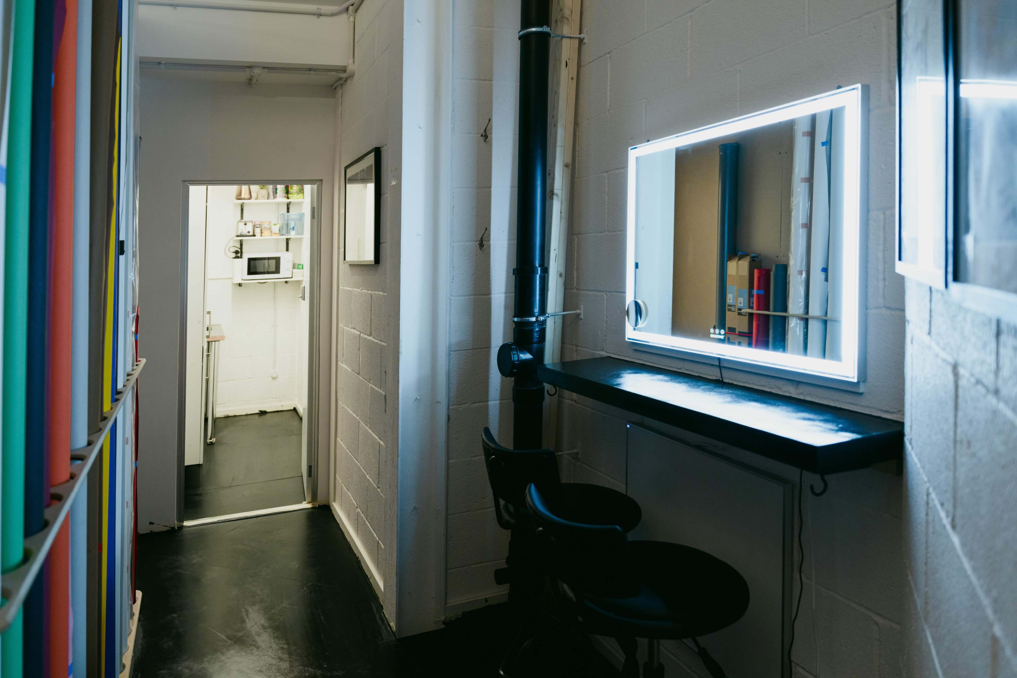 A narrow hallway features a brightly lit mirror and two stools alongside a white wall, with an open door leading to a kitchen area in the background.