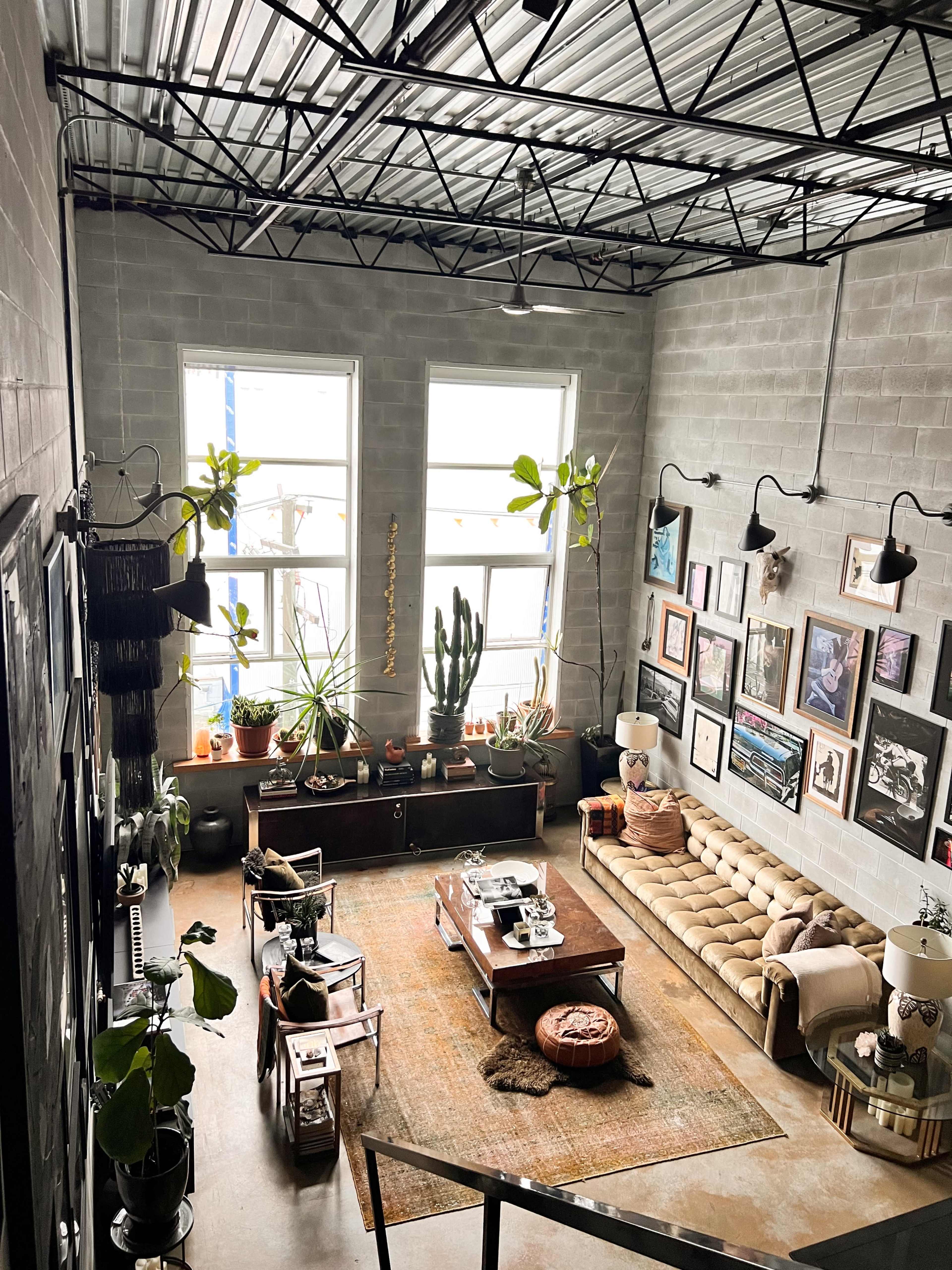 A spacious living room with large windows, a tan upholstered sofa, various plants, framed artwork on the walls, and a mix of modern and vintage furniture.