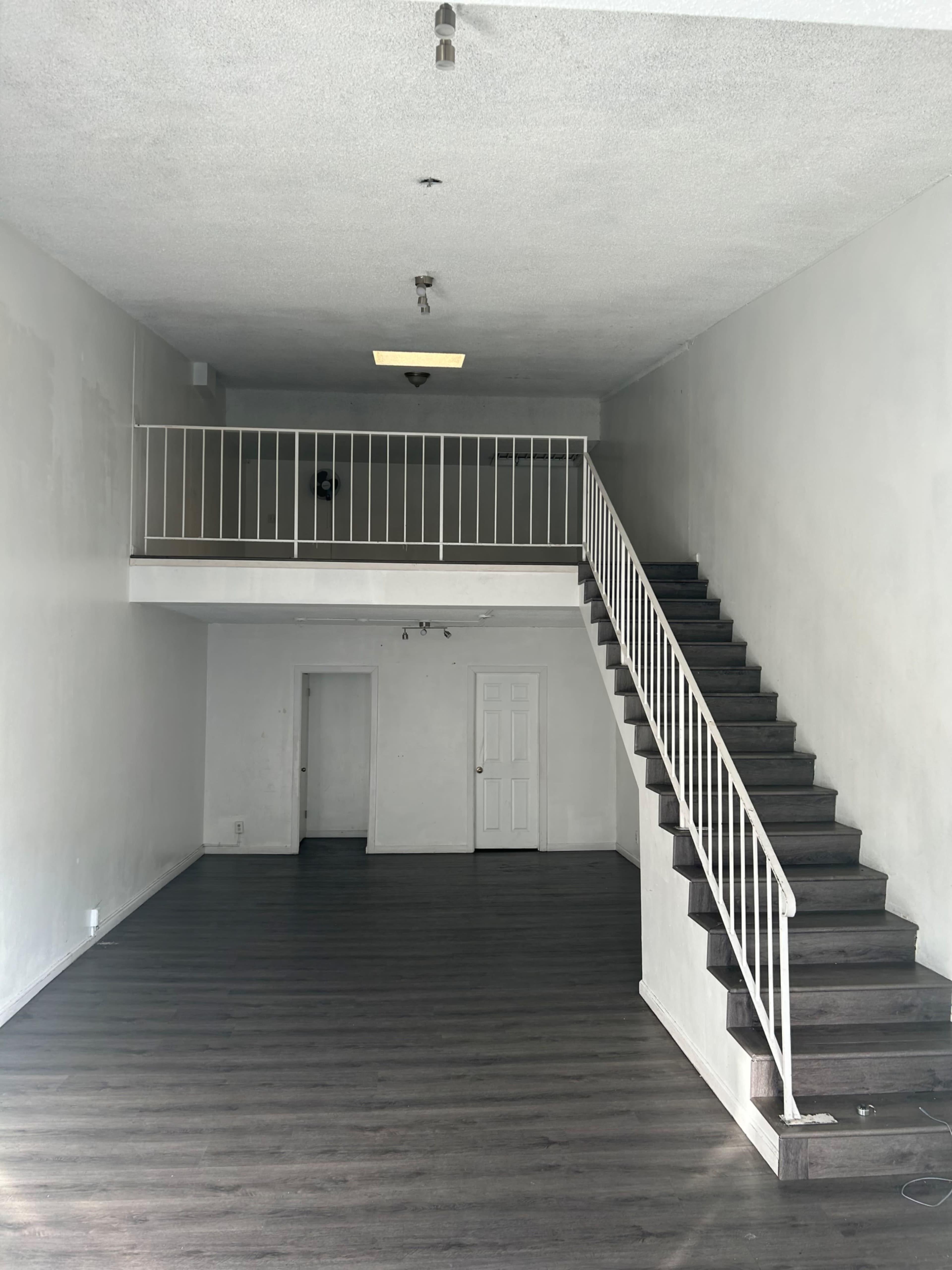 The image shows an empty room with a staircase leading to a small upper level, featuring a door on the left and white walls with a wooden floor.
