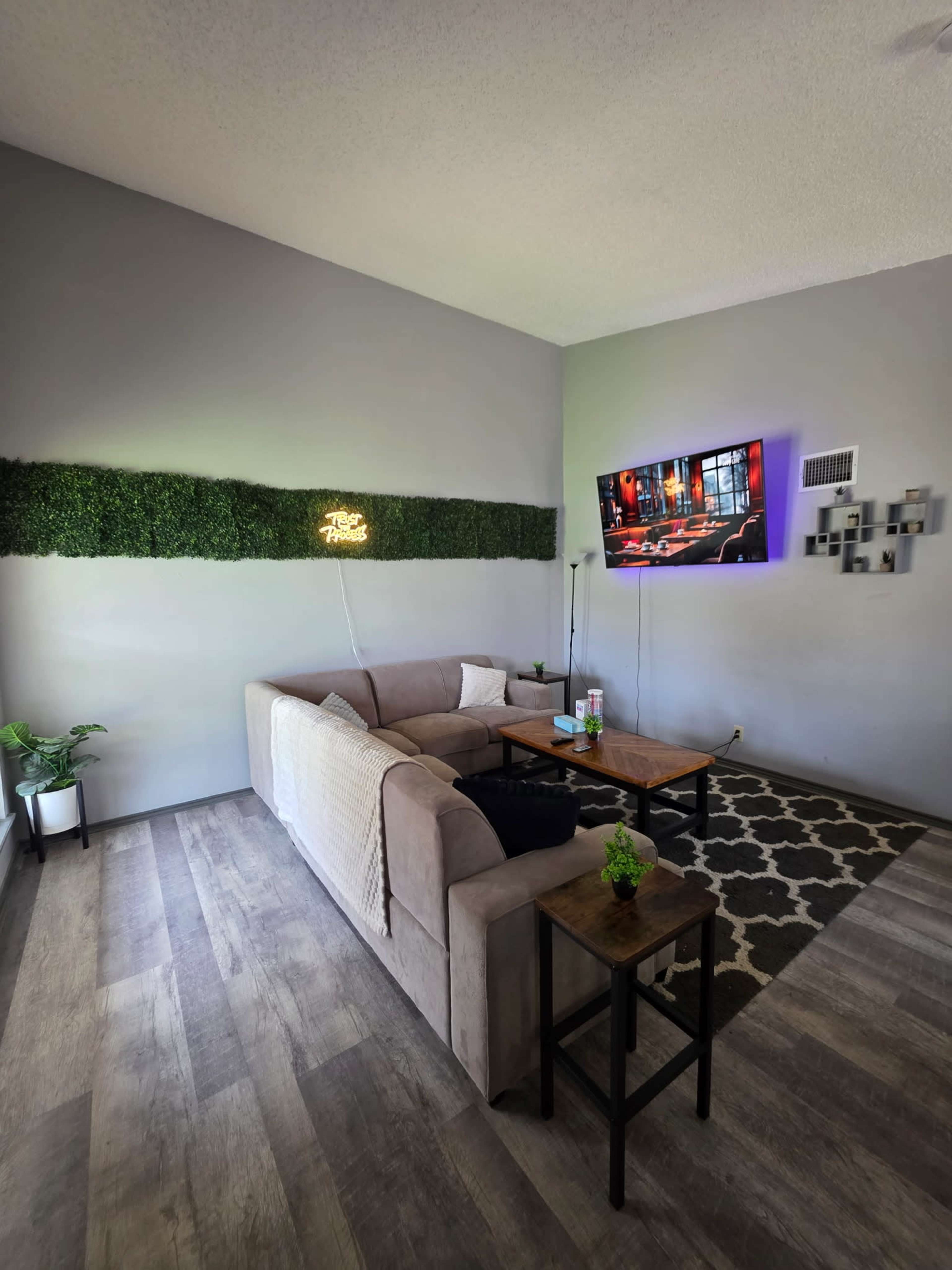 The image shows a modern living room with a beige couch, two wooden side tables, a television mounted on the wall, and a decorative green wall panel.