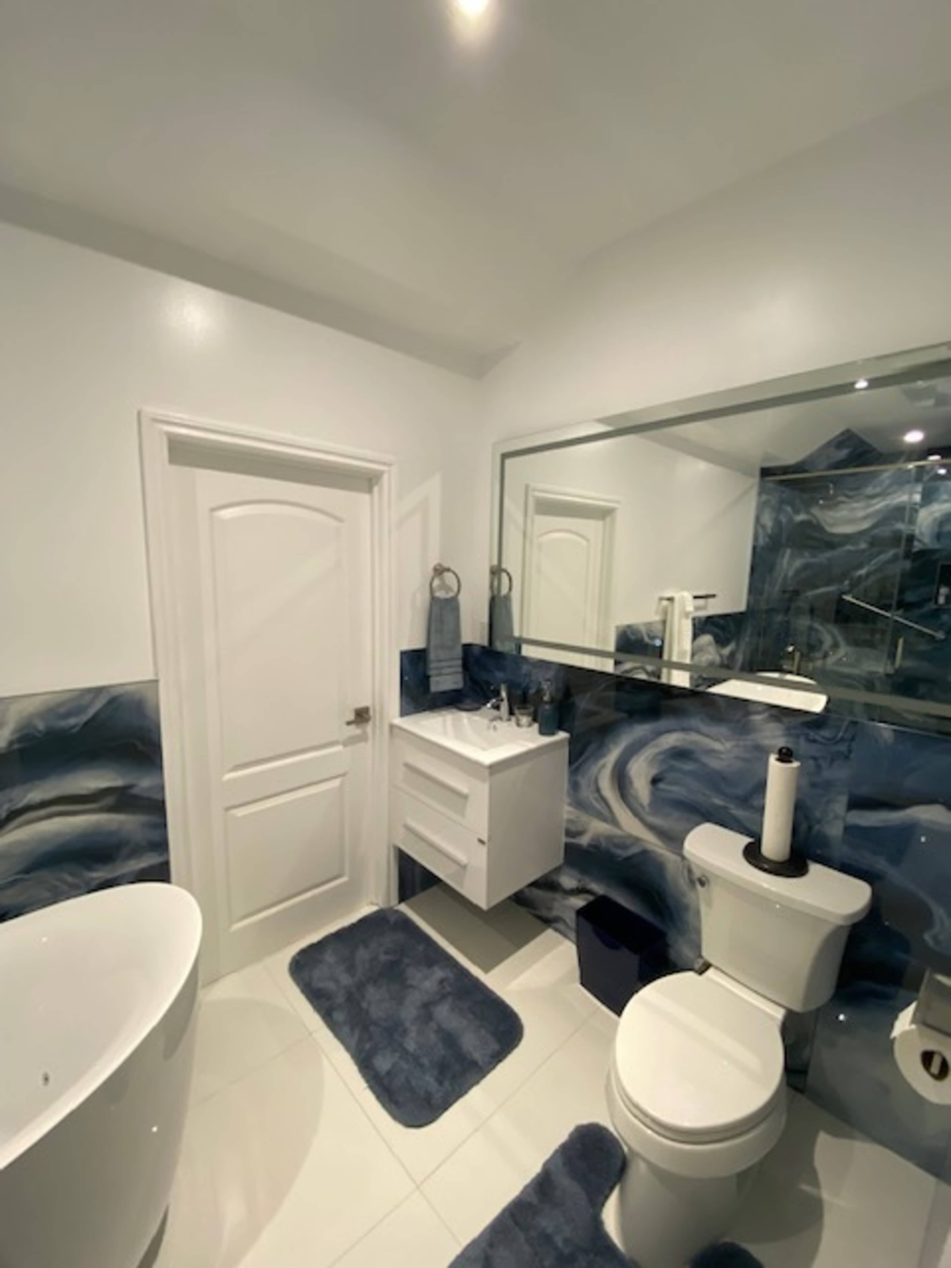 The bathroom features a modern design with a freestanding tub, a wall-mounted vanity, a toilet, and a large mirror, all set against a blue and white marbled wall.