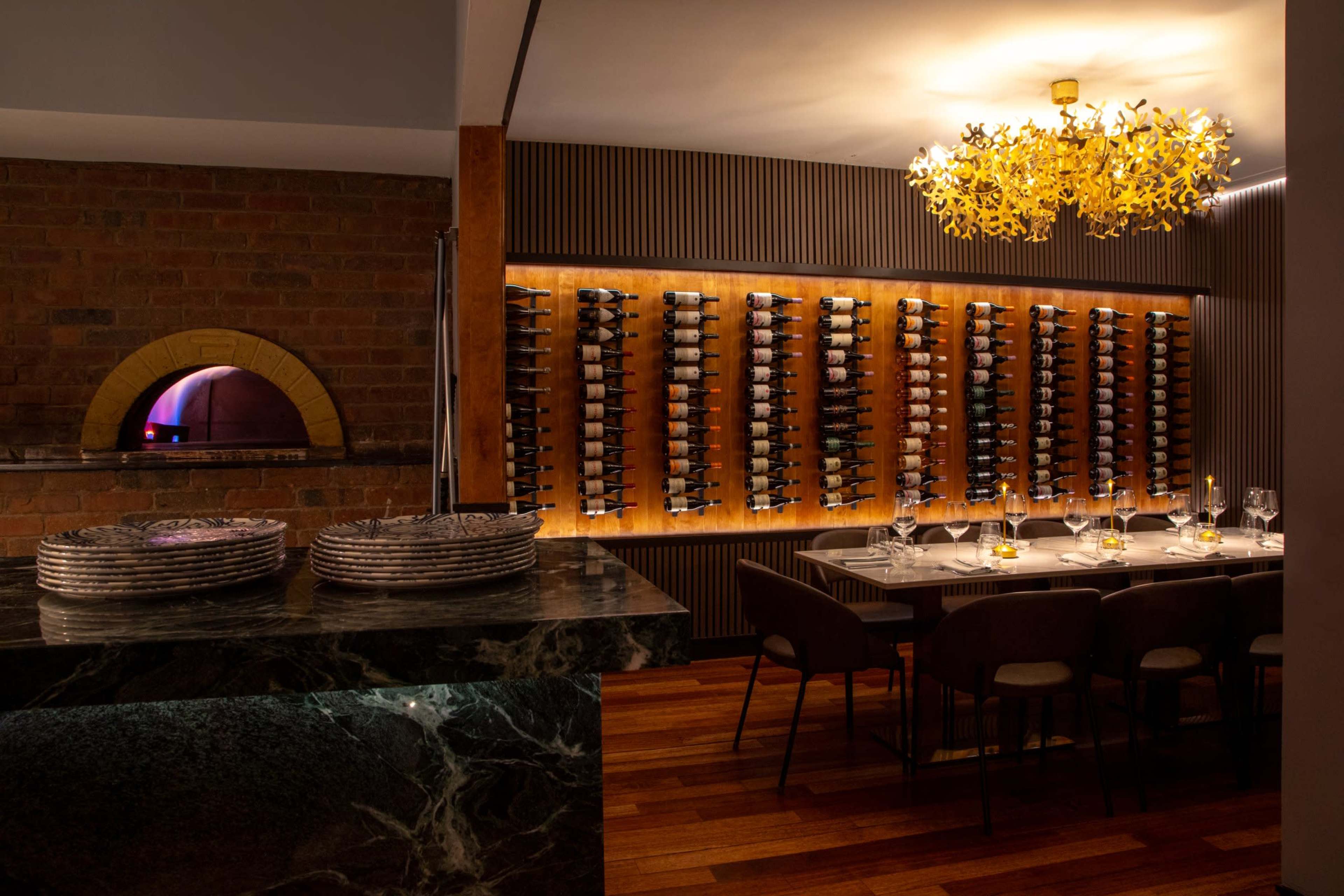 The image shows a modern dining area featuring a wine wall, a large wooden table set with dinnerware, and a stone pizza oven in the background.