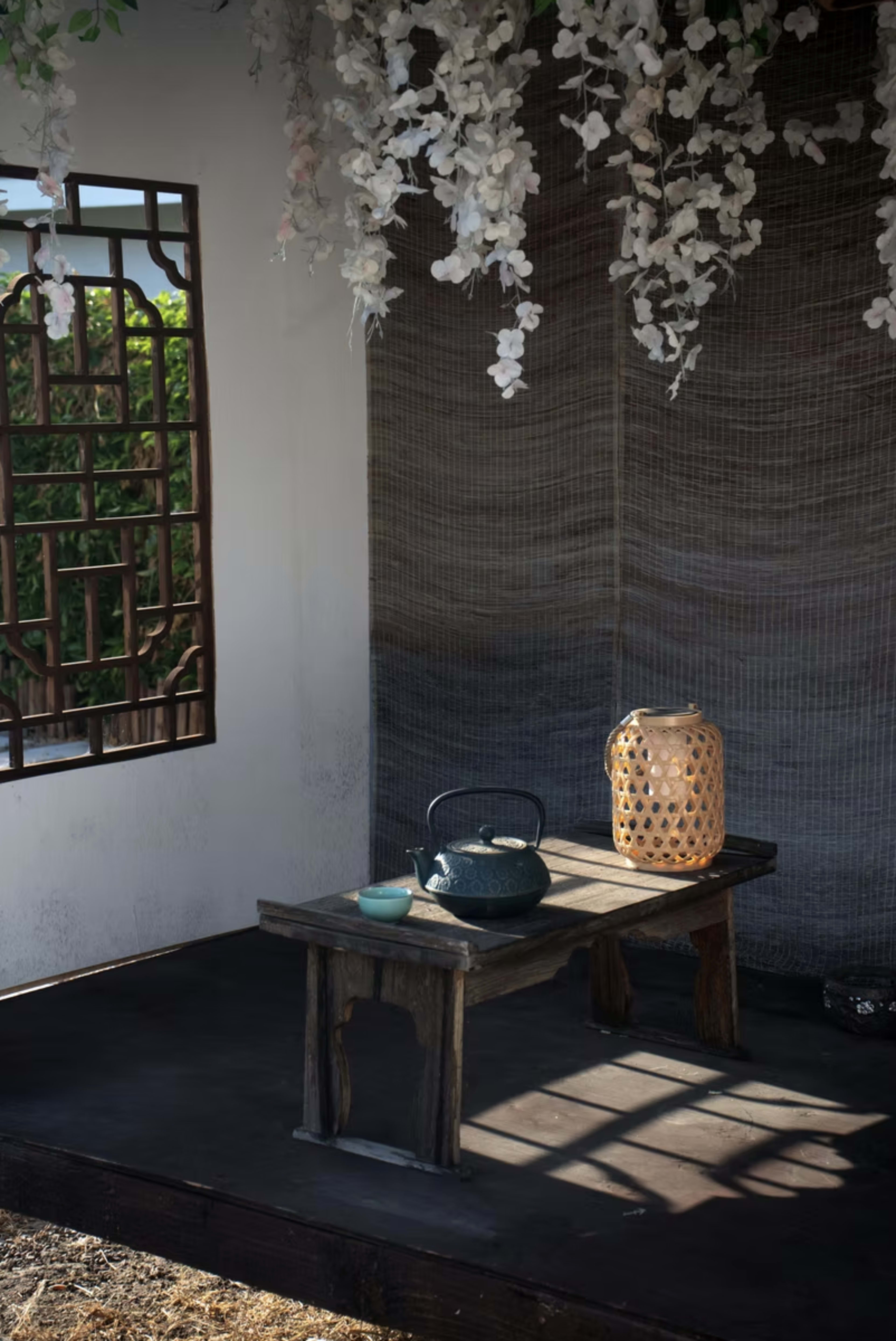 The image features a serene indoor space with a wooden table displaying a tea kettle and a decorative lantern, surrounded by flowering vines hanging from the ceiling.