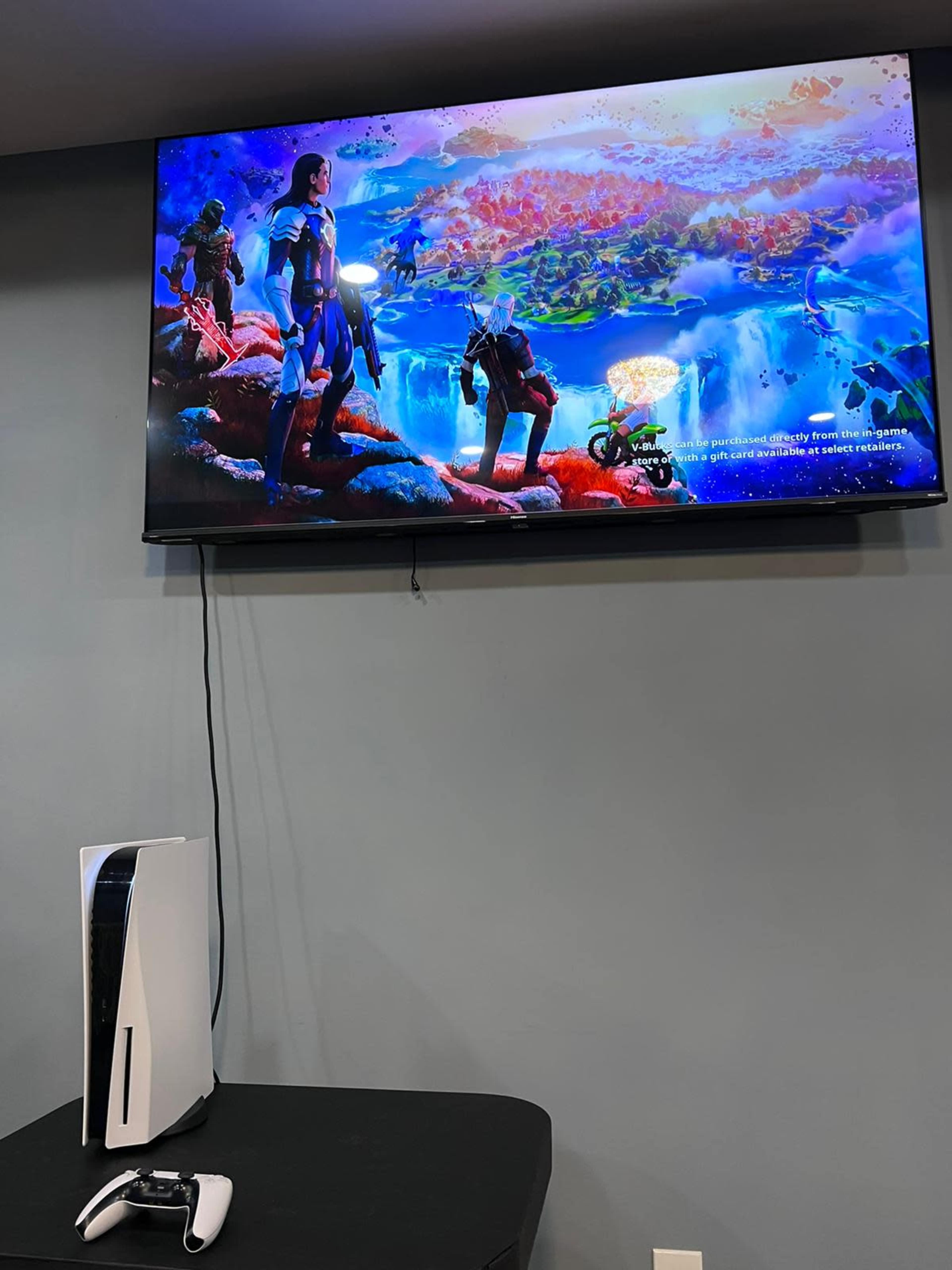 A PlayStation 5 console and controller sit on a table beneath a large TV displaying a vibrant video game scene.