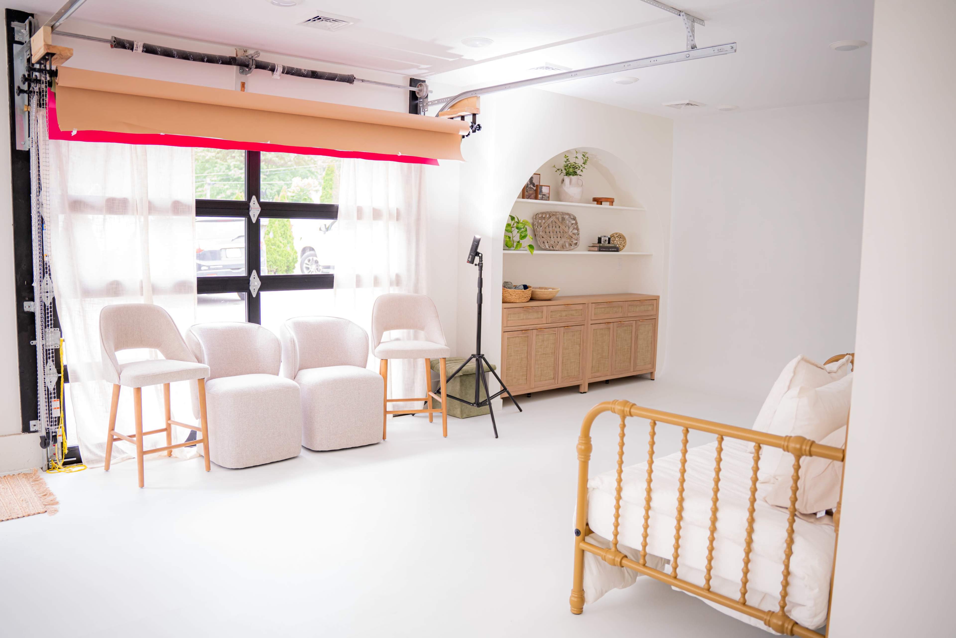 The image shows a bright, minimalist interior space with a seating area featuring three upholstered chairs, a wooden cabinet, and a bed against the wall.