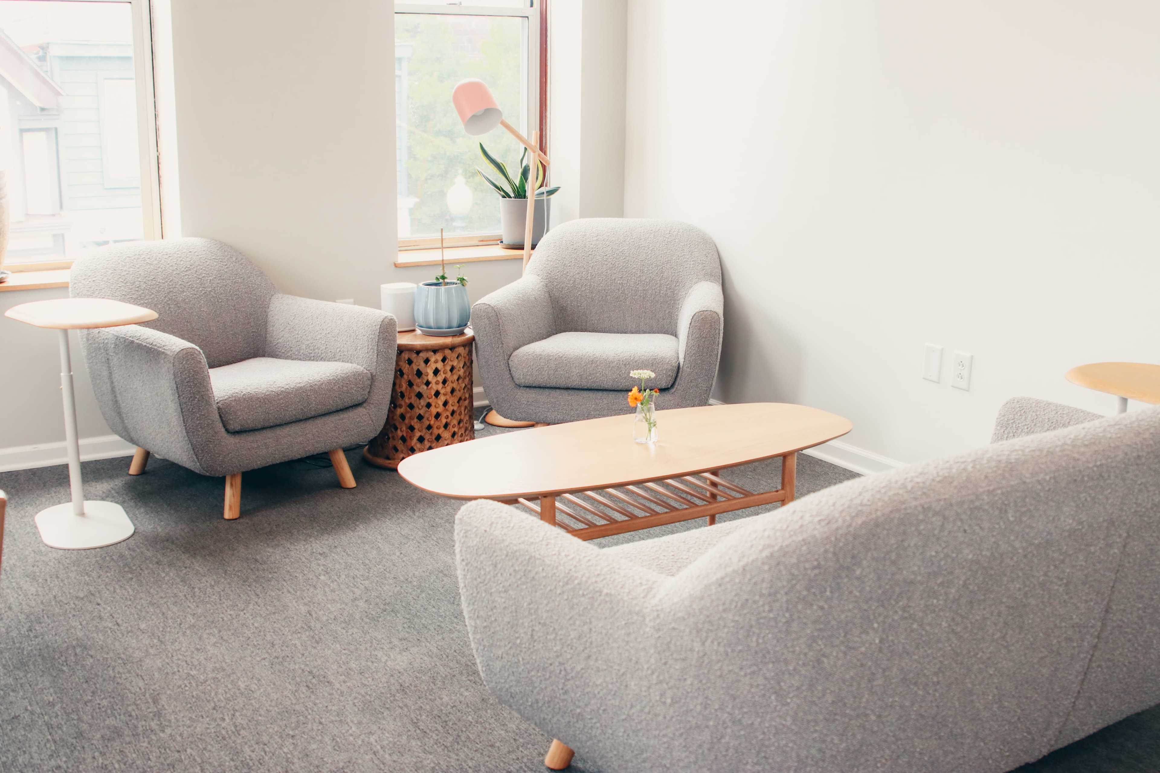 The image shows a cozy seating area with two gray armchairs, a small wooden coffee table, and a decorative stool, illuminated by natural light from nearby windows.
