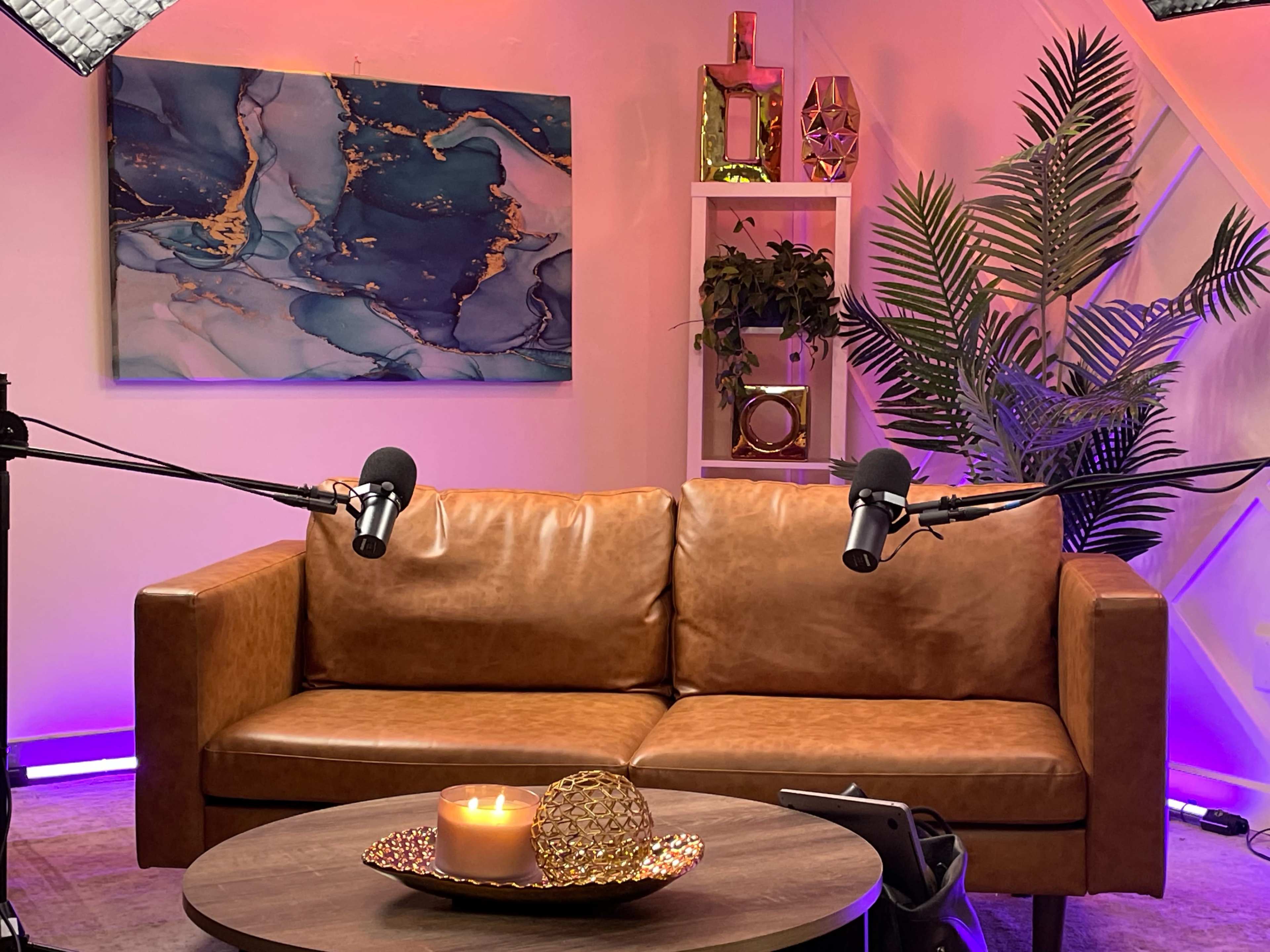 The image shows a cozy studio setup featuring a brown leather couch with two microphones positioned in front of it, alongside a candle and decorative items on a round table, with colorful lighting and a decorative wall in the background.