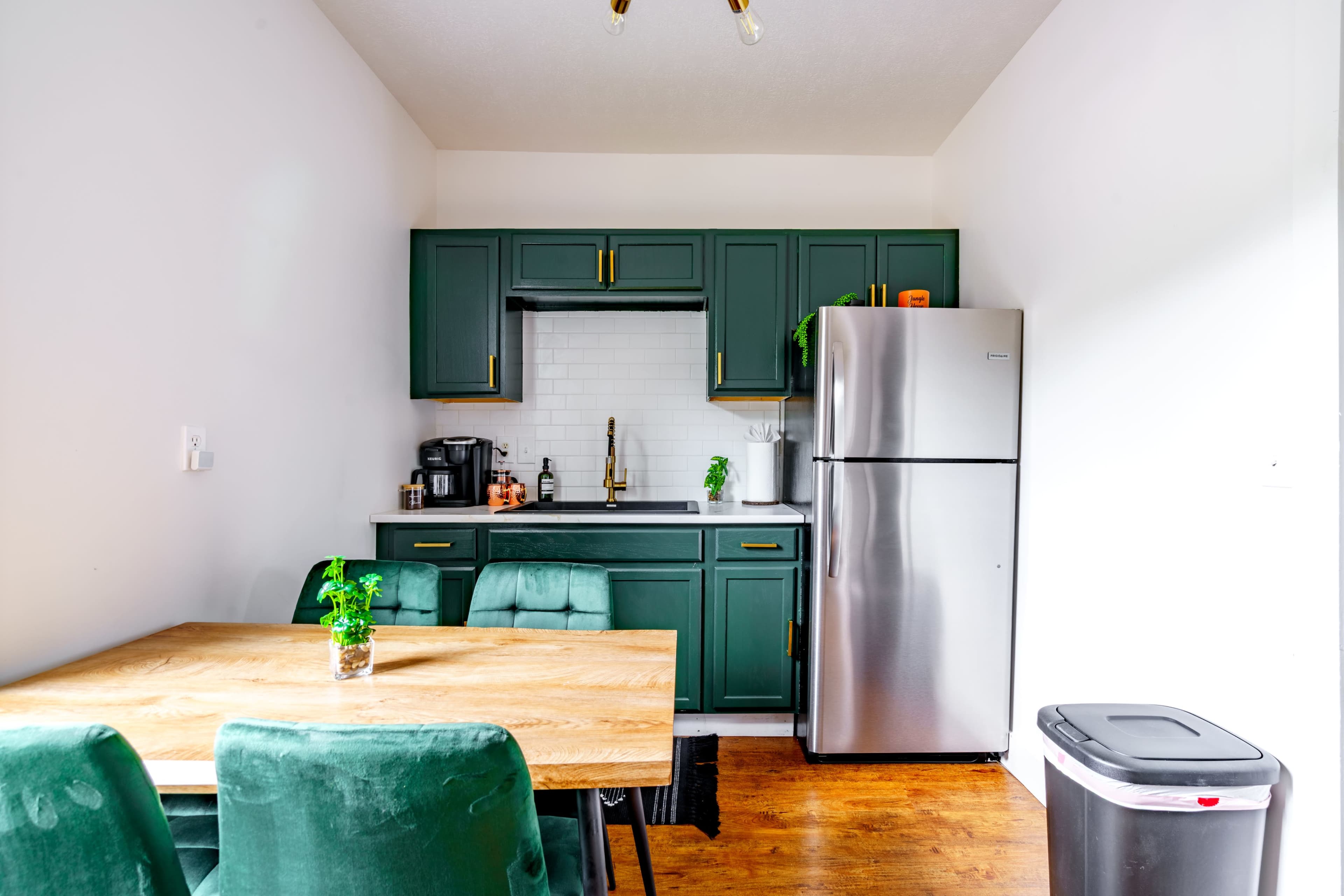 A modern kitchen features green cabinets, a stainless steel refrigerator, and a wooden dining table with green chairs.