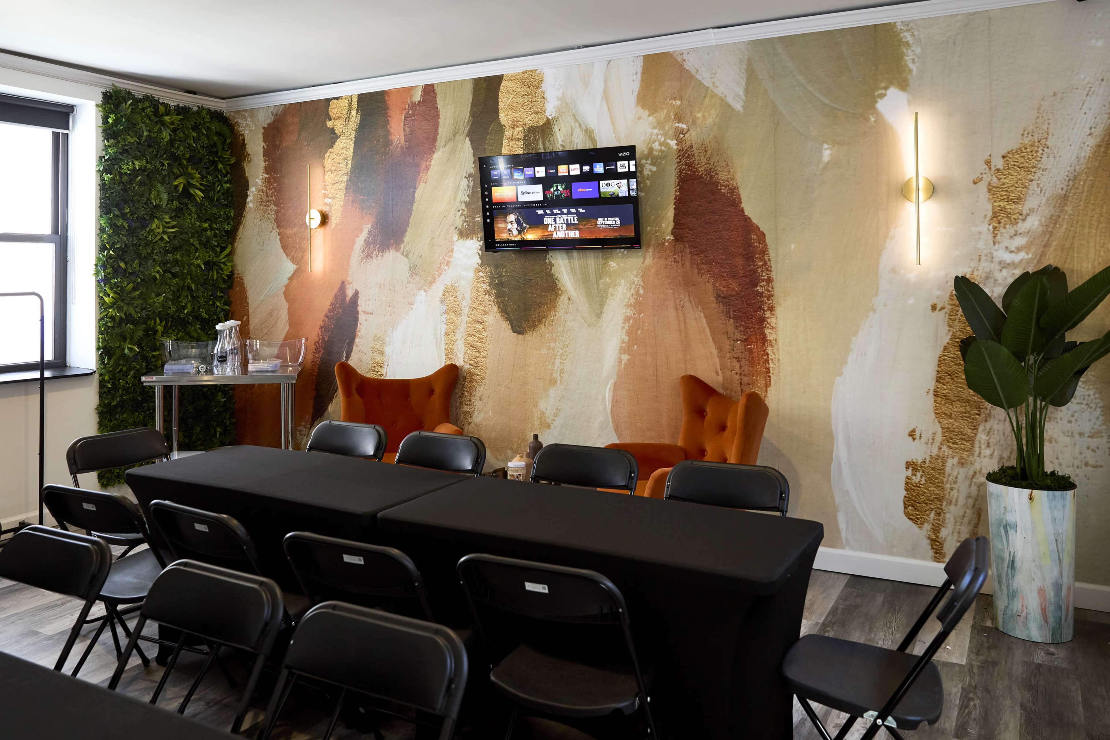 The image shows a modern conference room with a large patterned wall, black tables arranged in rows, and orange accent chairs.