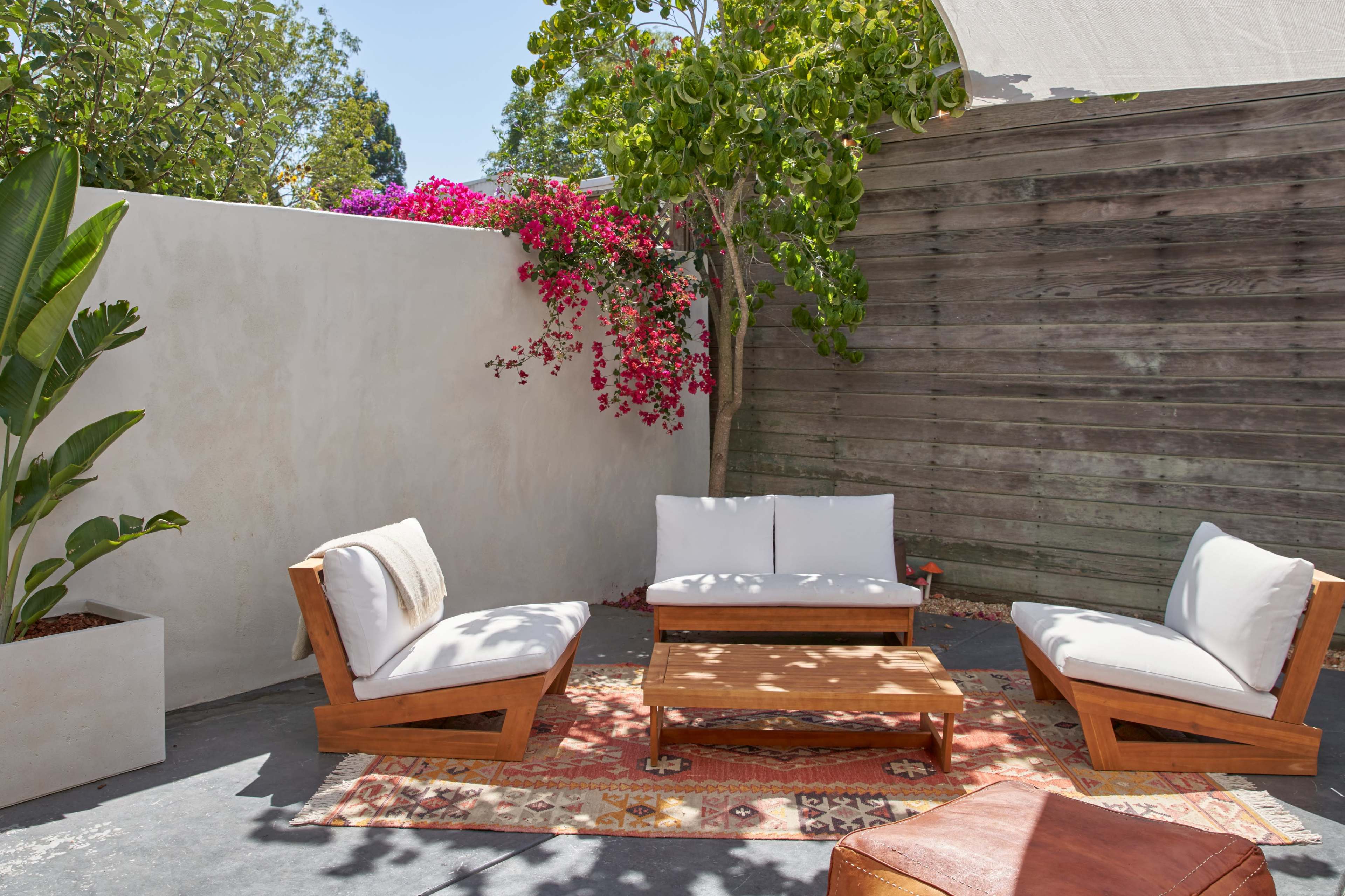 A cozy outdoor seating area features two wooden chairs and a low table on a patterned rug, surrounded by lush greenery and a wall adorned with pink bougainvillea.