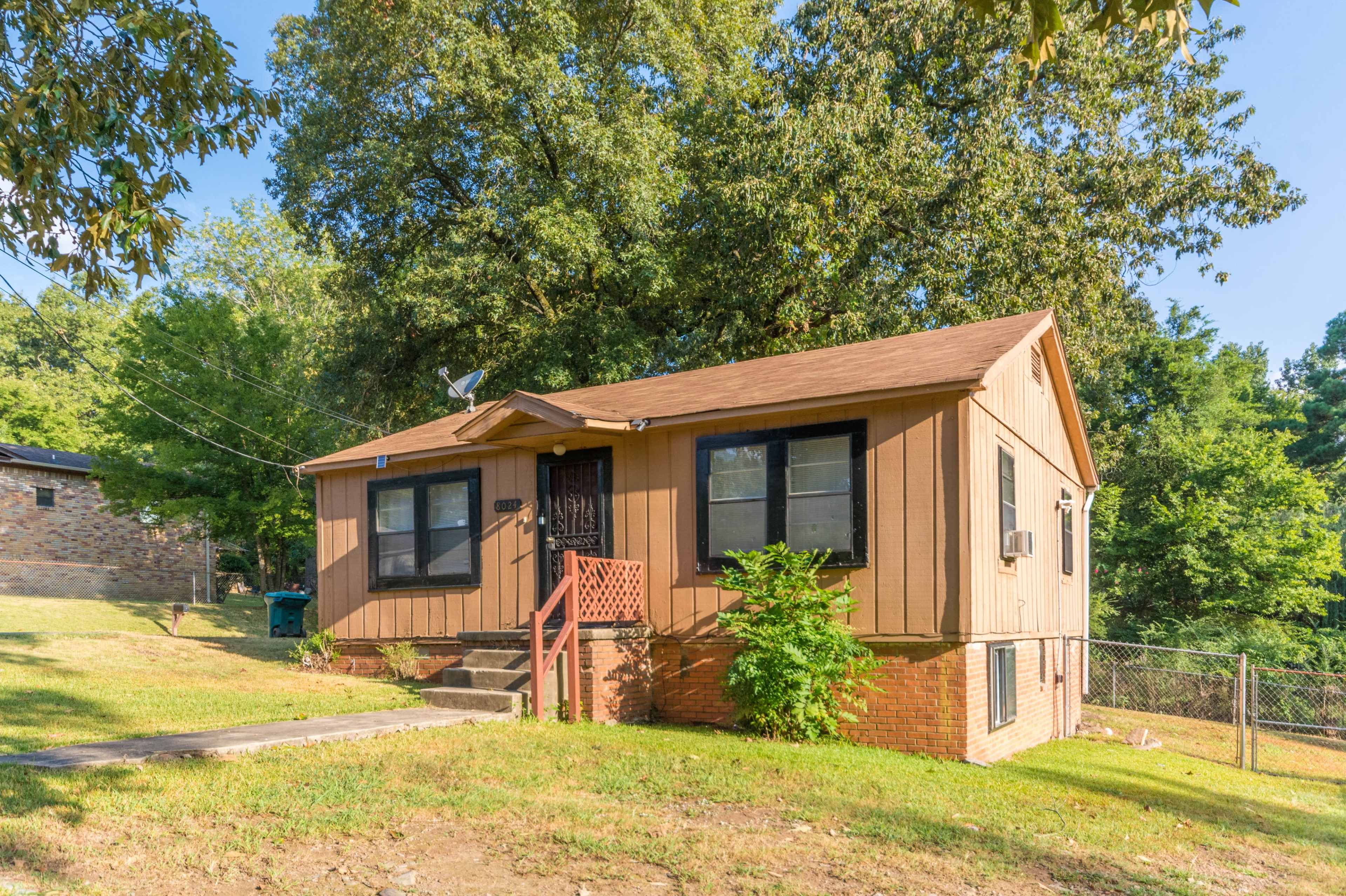 A small, single-story house with a brown wooden exterior, located on a grassy lot surrounded by trees.