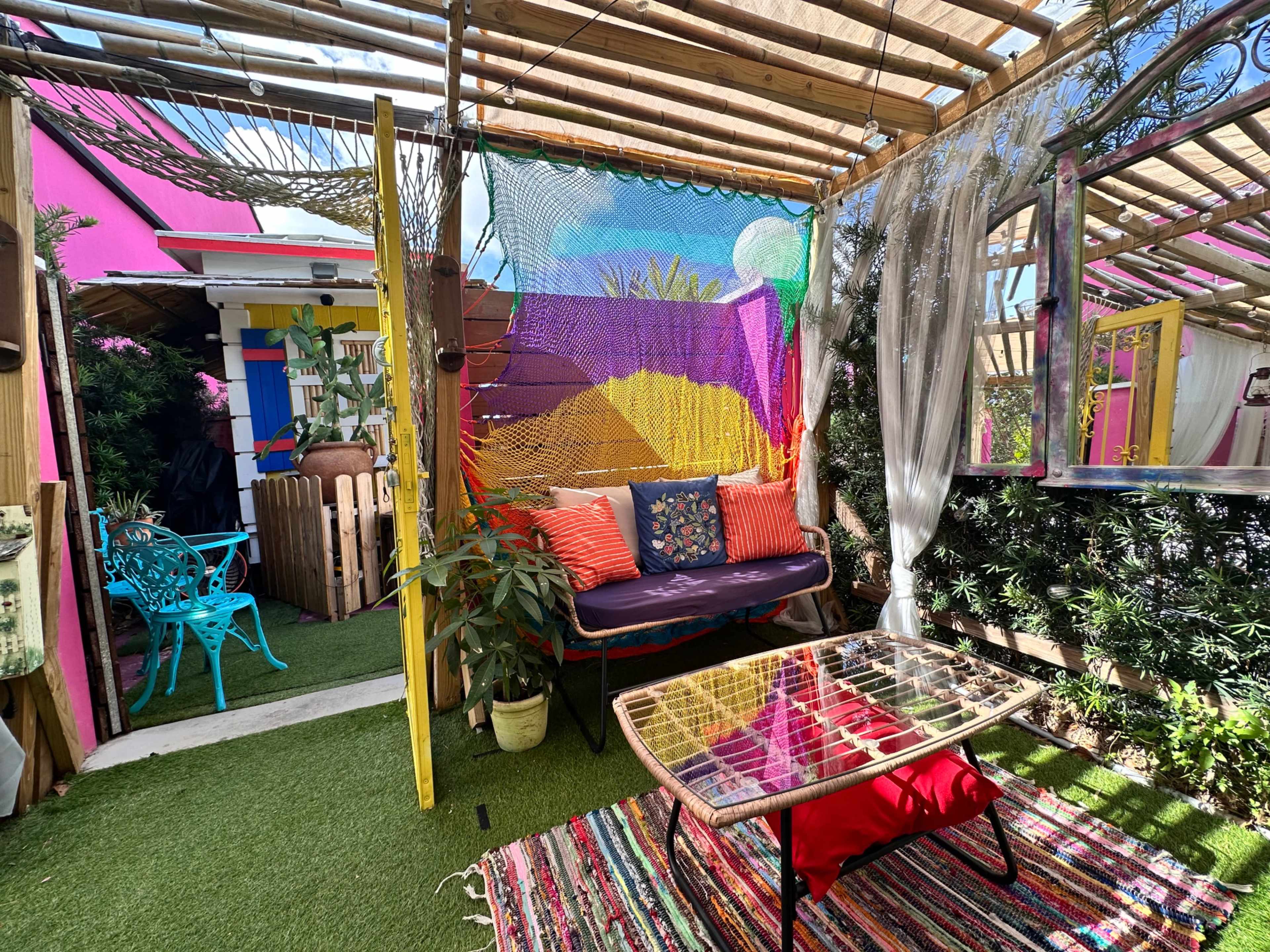 The image shows a colorful outdoor seating area with a couch adorned with cushions, a small table, and vibrant decorations, surrounded by greenery and an intricate canopy.