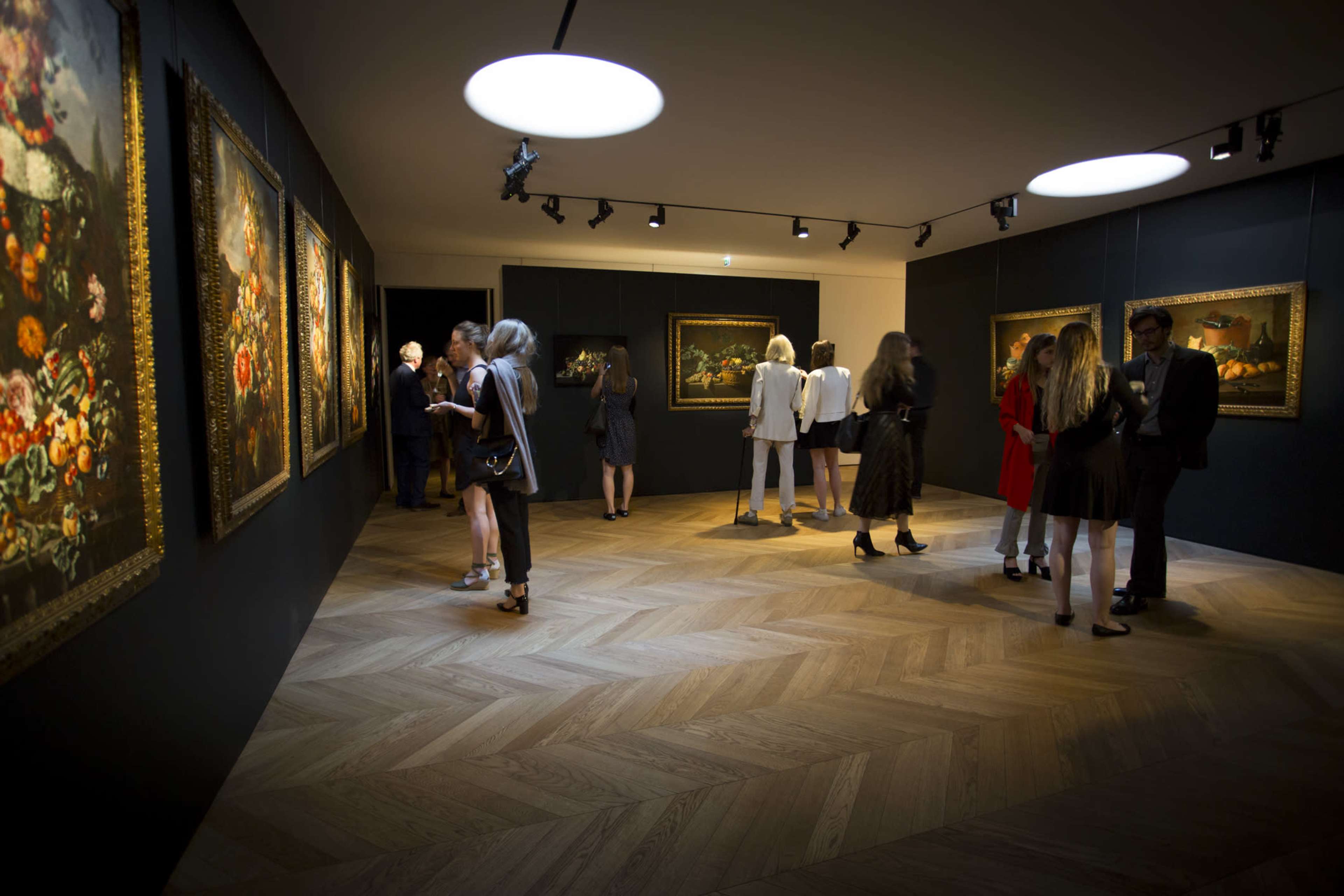 A group of people explores an art gallery featuring framed paintings on dark walls and wooden flooring.