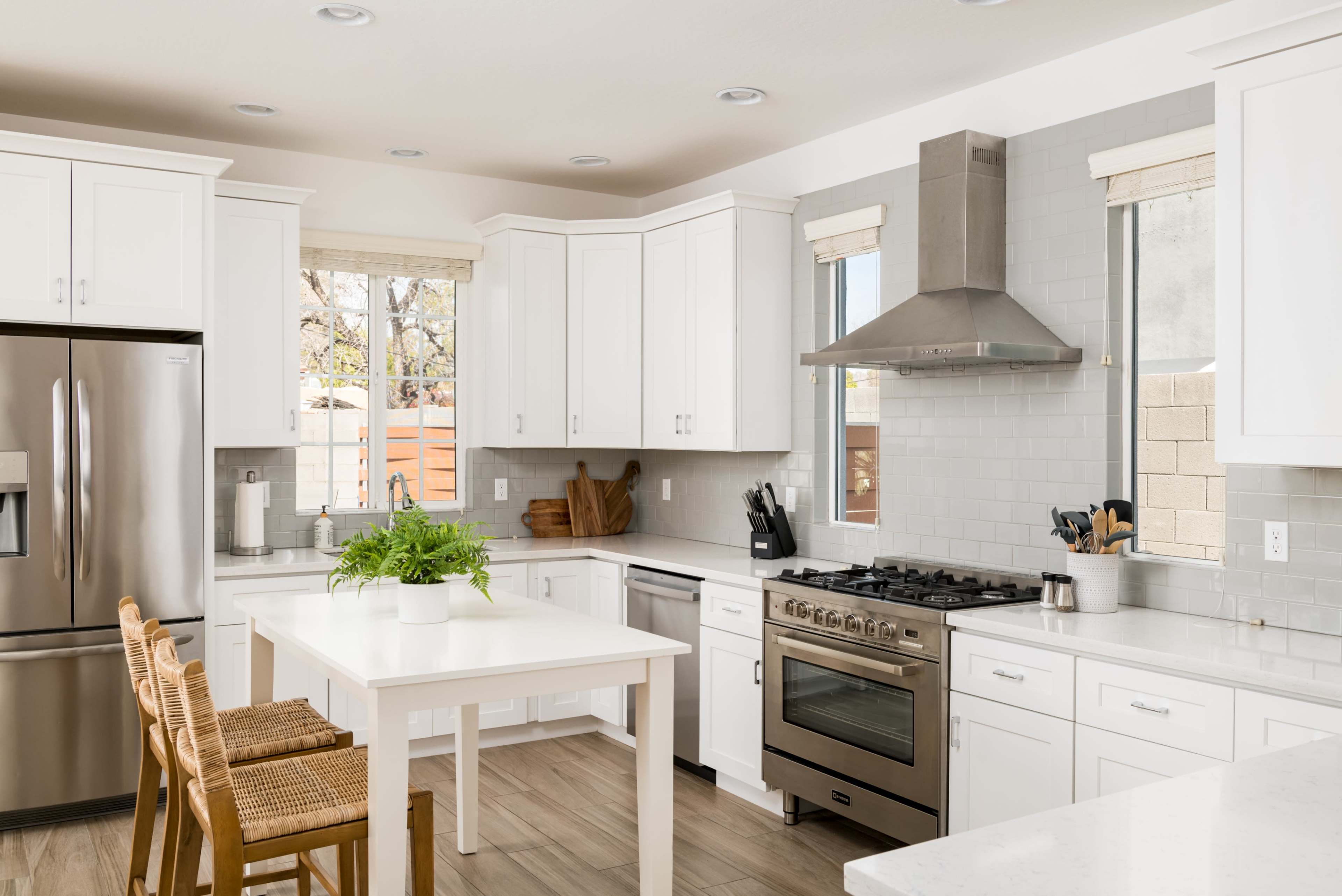A modern kitchen features white cabinetry, stainless steel appliances, and a central table with barstools and a potted plant.
