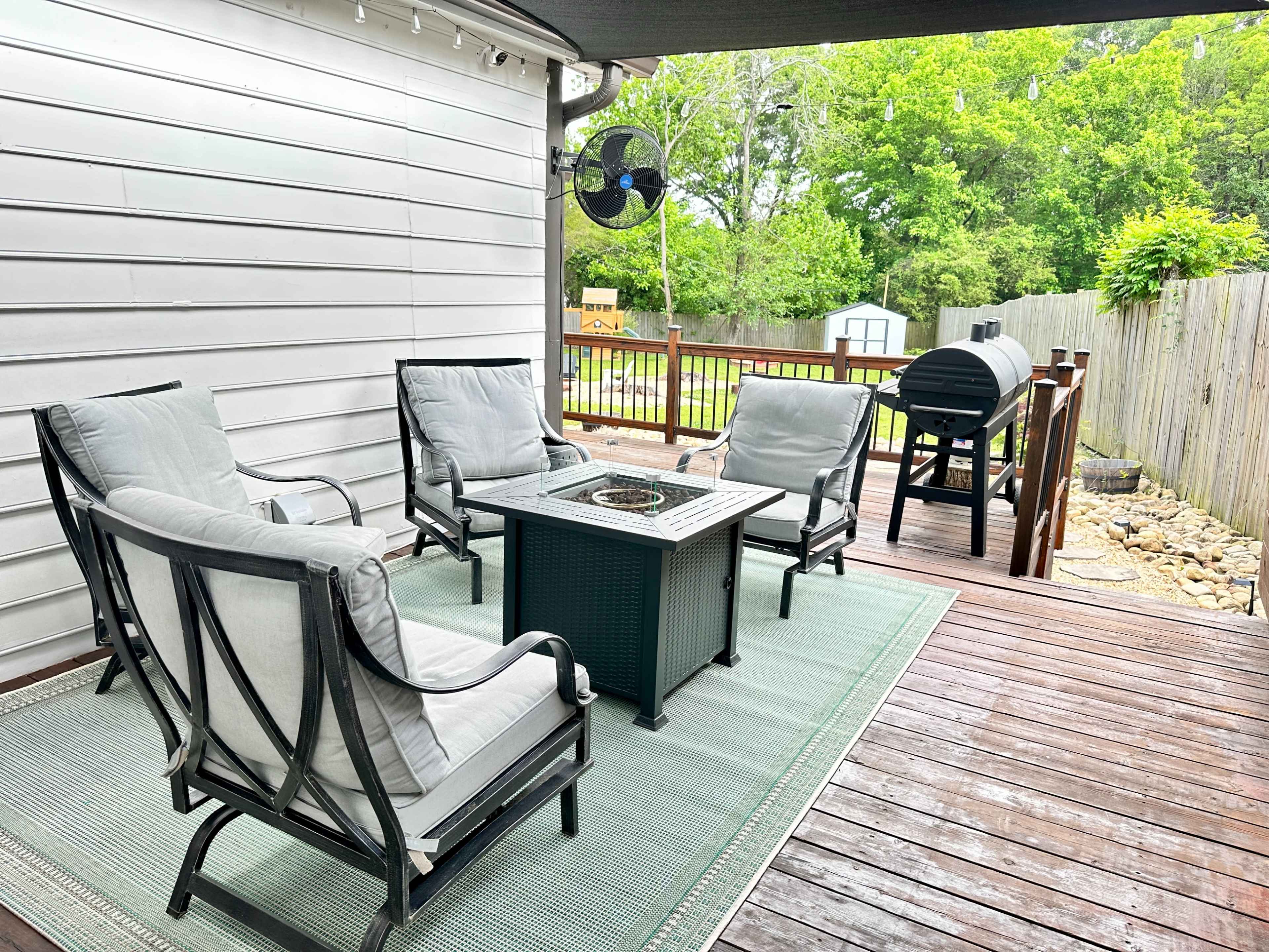 A patio area features four chairs arranged around a fire pit table on a wooden deck, with a grill and greenery visible in the background.
