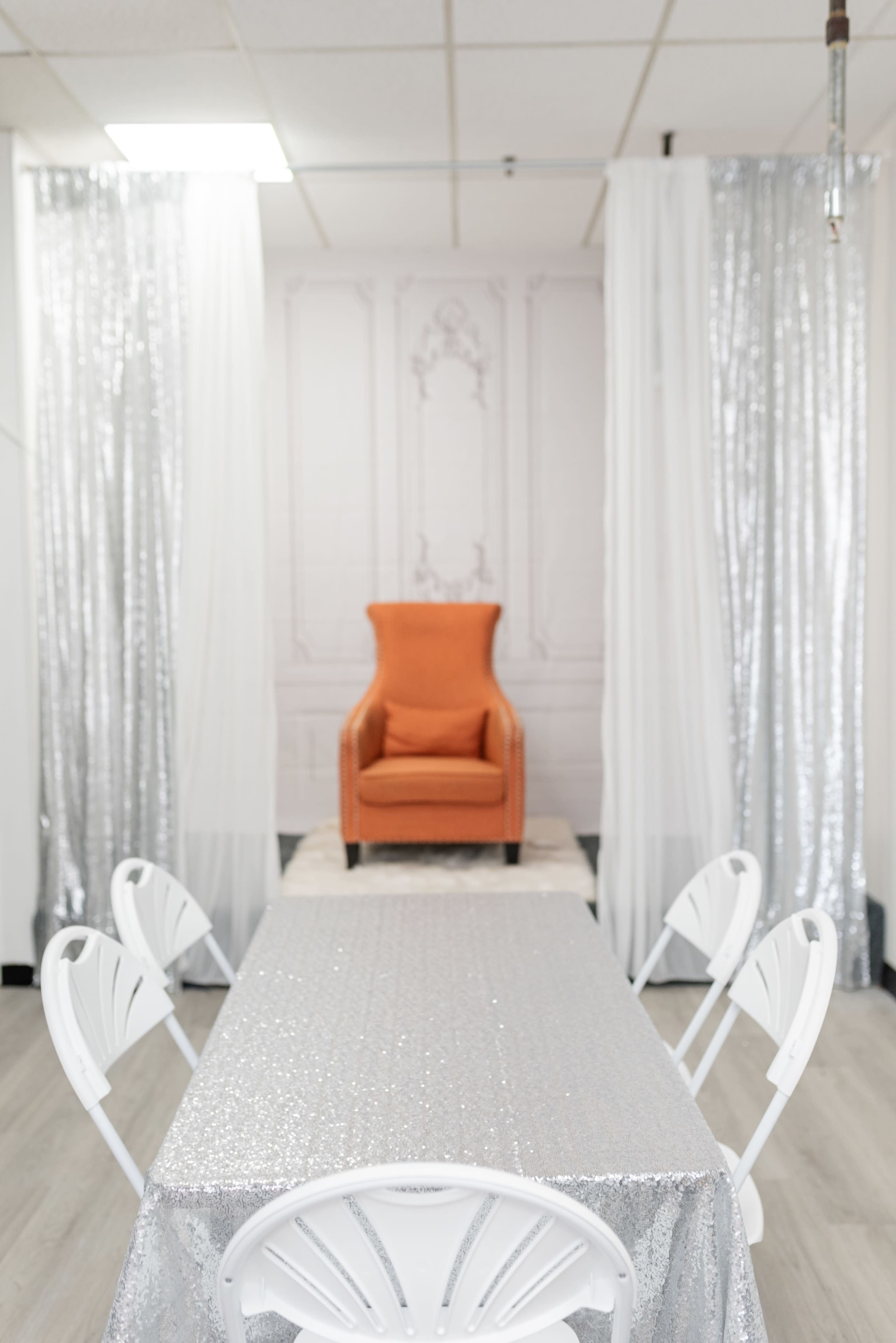 The scene features a well-lit room with a sparkling silver table and an orange armchair at the back, flanked by sheer white and silver drapes.