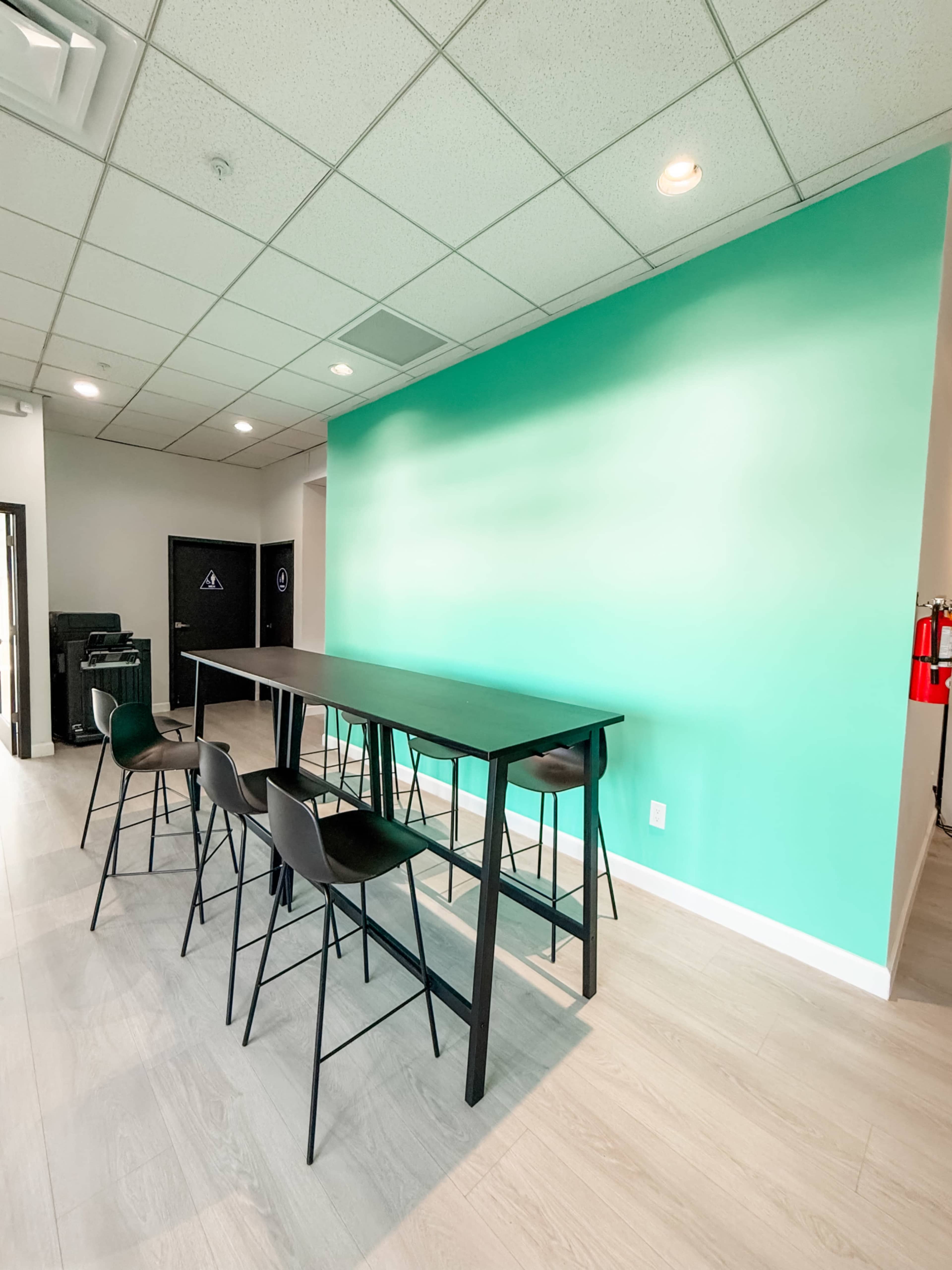 The image shows a modern workspace featuring a black table with high chairs against a vibrant teal wall.
